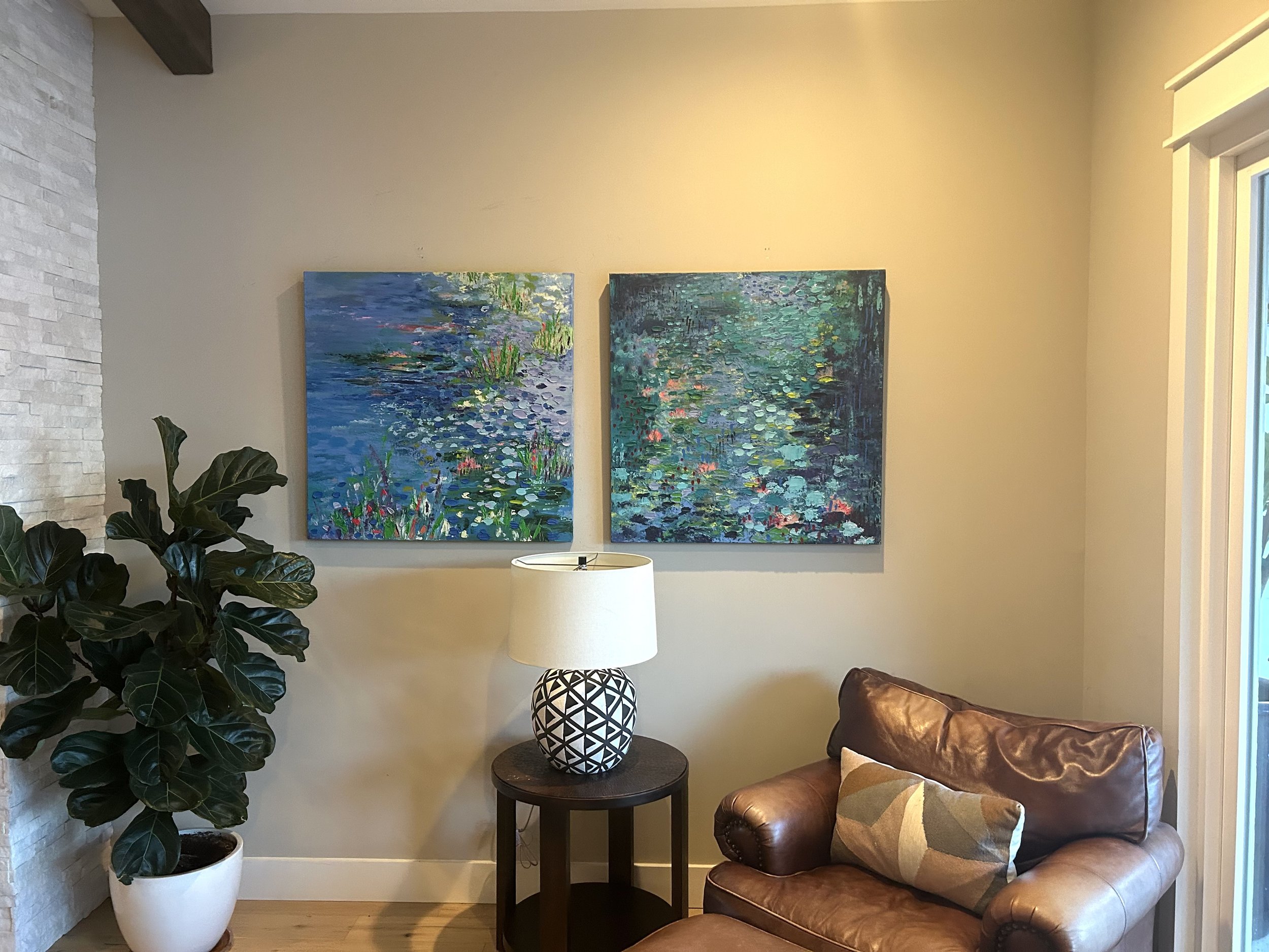 Living room corner with two abstract water lily paintings on beige wall, a large potted plant on the left, a black side table with a geometrically patterned lamp in the center, and a brown leather armchair with a decorative pillow on the right.