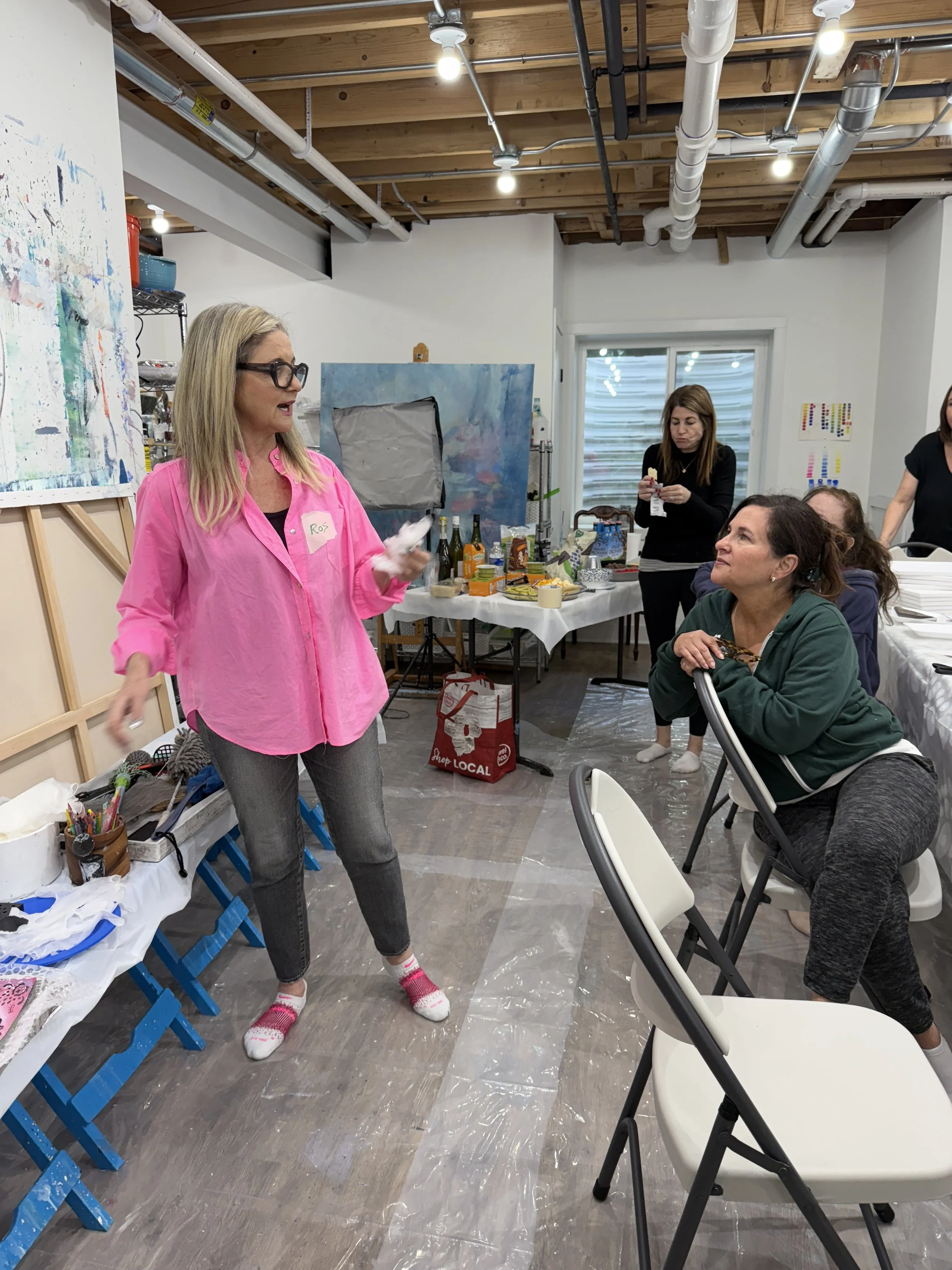 A woman in a pink shirt with glasses is standing and speaking to a seated woman in a green hoodie who is listening attentively. Two other women are in the background, one standing and the other partially visible. The setting appears to be an art studio or craft room, with supplies on a table, artwork on the walls, and a wooden ceiling with exposed beams and pipes. The table is covered with various food and drink items.