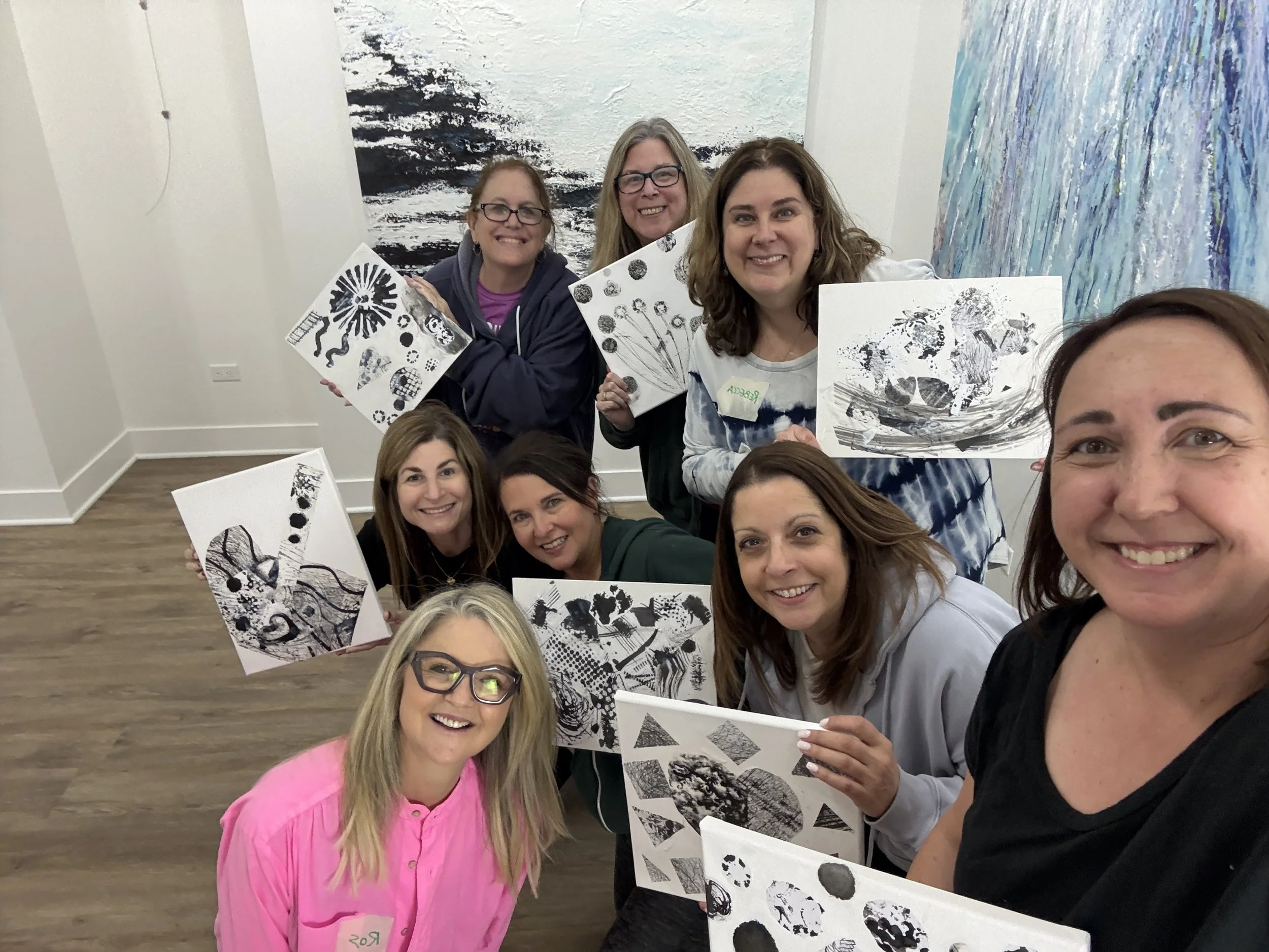 A group of smiling women posing together, holding black and white abstract art pieces, in an indoor setting with white walls and colorful paintings in the background.