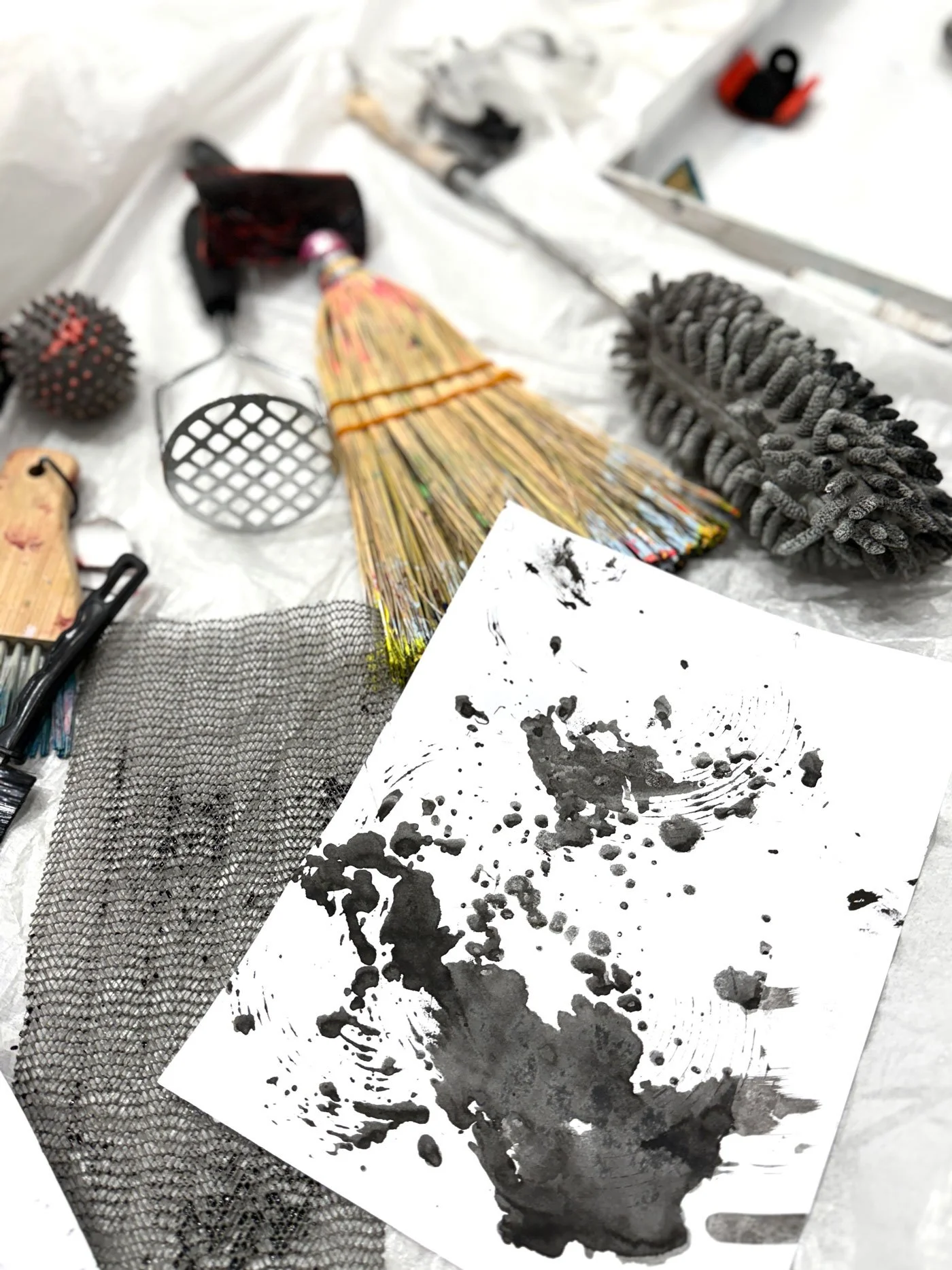 A flat surface with various objects including an abstract black ink splatter on a white sheet of paper, a black mesh material, a bundle of colorful sticks, a fuzzy gray object, a small round spiky object, a metal grate, a small knife, and other miscellaneous items.
