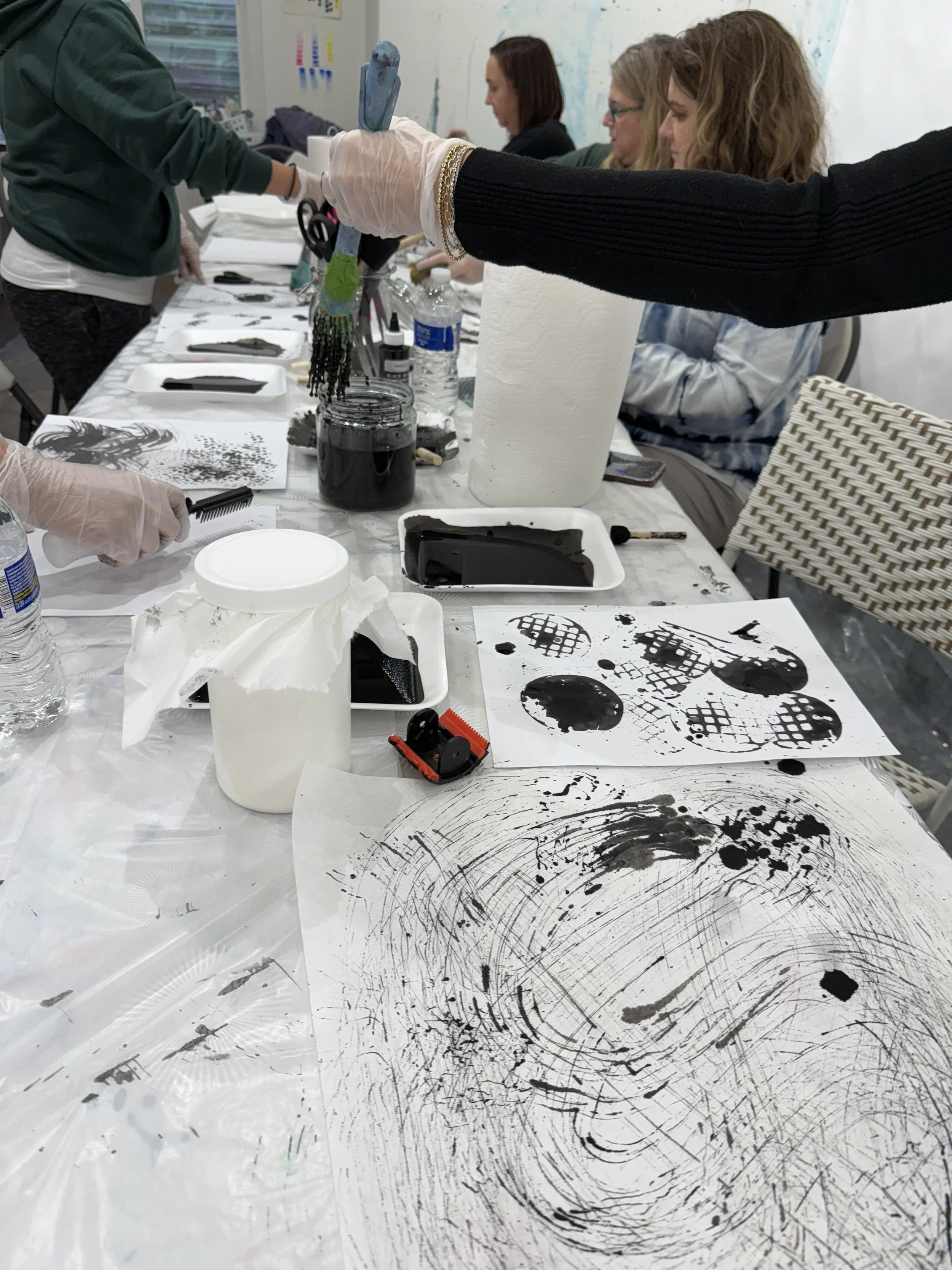 People at a table participating in a black ink painting or printing activity, with ink, brushes, and paper scattered on the table.
