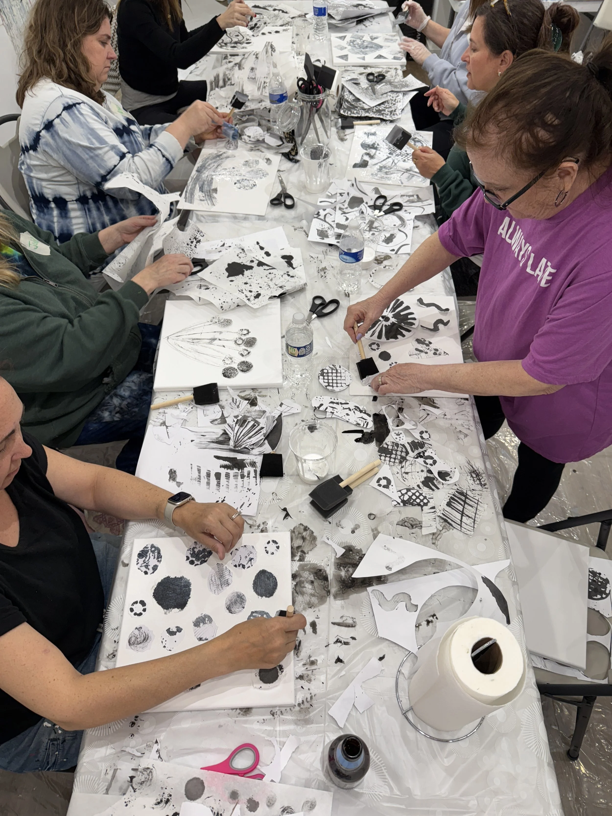 People participating in a paper arts or printmaking workshop at a table covered with black and white painted paper, scissors, and art supplies.