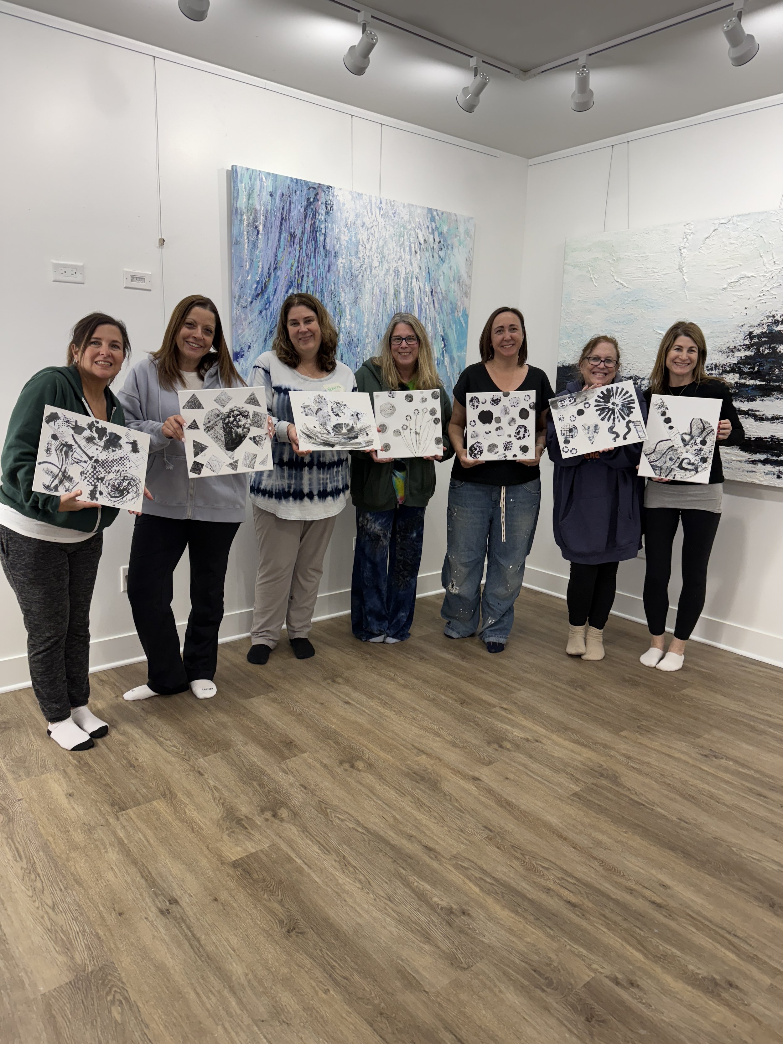 Group of seven women standing in an art gallery, each holding a black-and-white abstract artwork, with large textured paintings on the wall behind them.