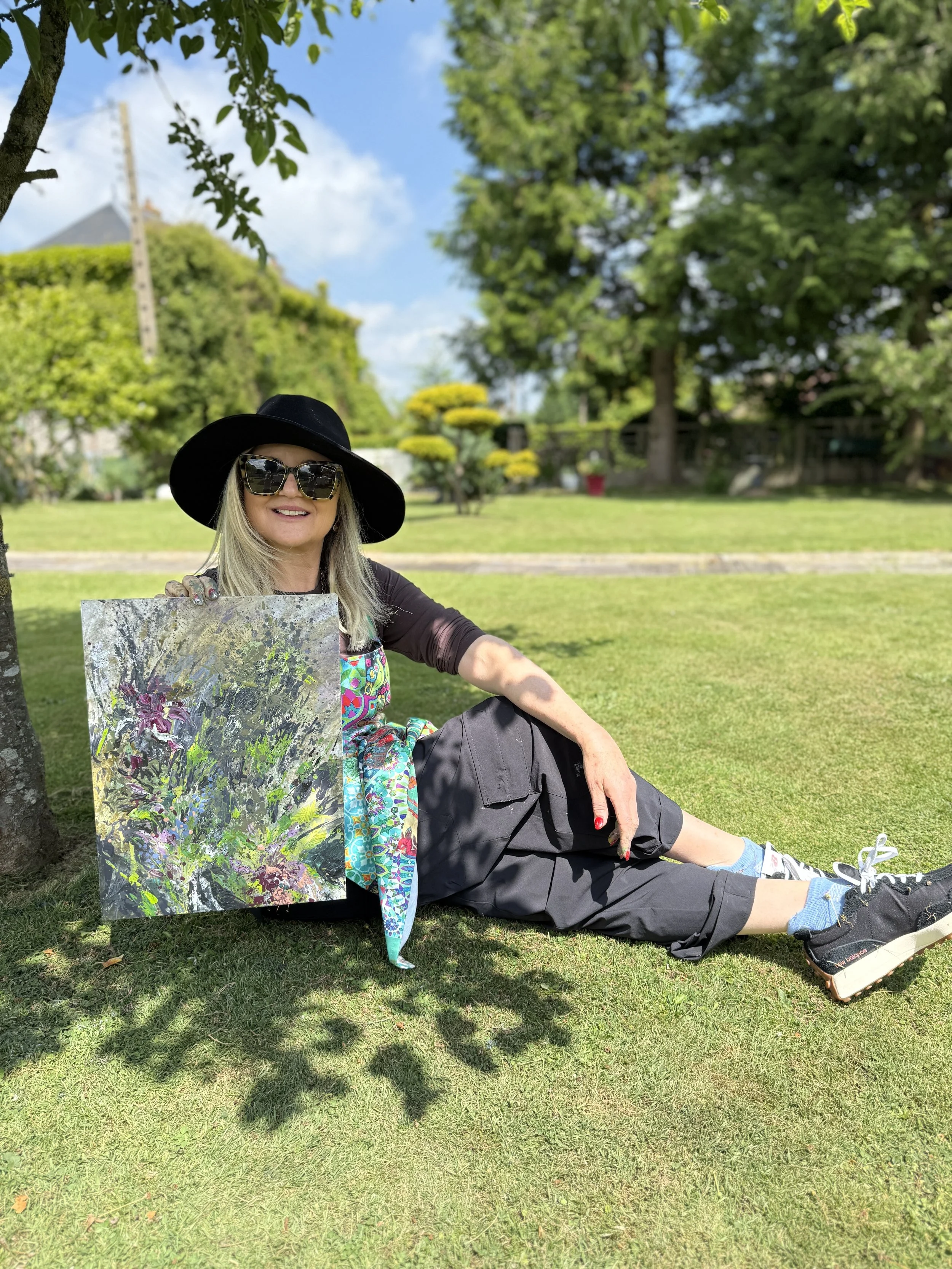 A woman wearing sunglasses and a black hat sitting on grass in a park, holding a colorful abstract painting, with trees and a clear blue sky in the background.