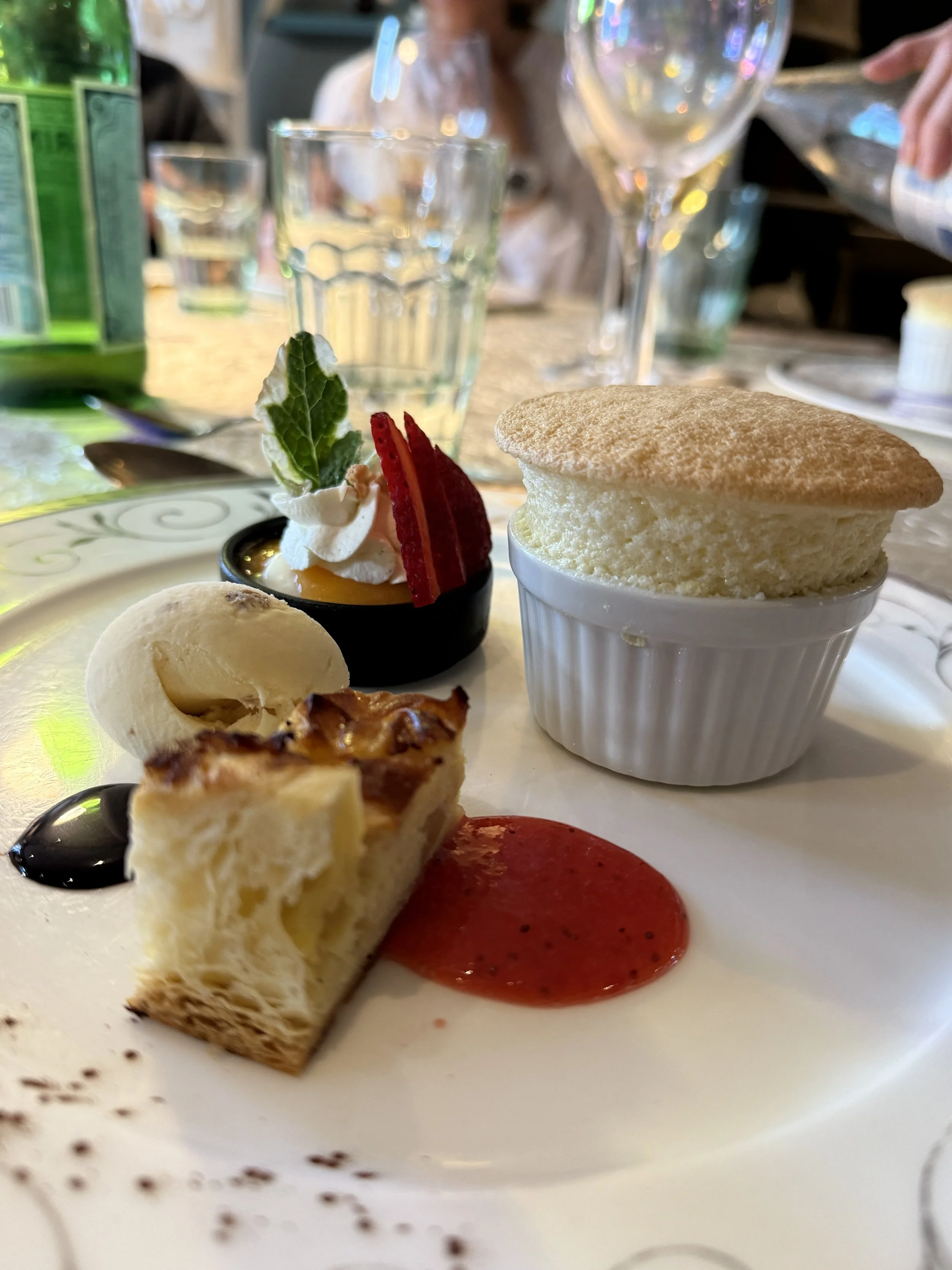 Close-up of a dessert plate featuring a slice of cheesecake, a small tart with whipped cream, a strawberry, a sprig of mint, and a dollop of red sauce on a white plate. In the background, there are glasses of water and wine, and a blurry person.