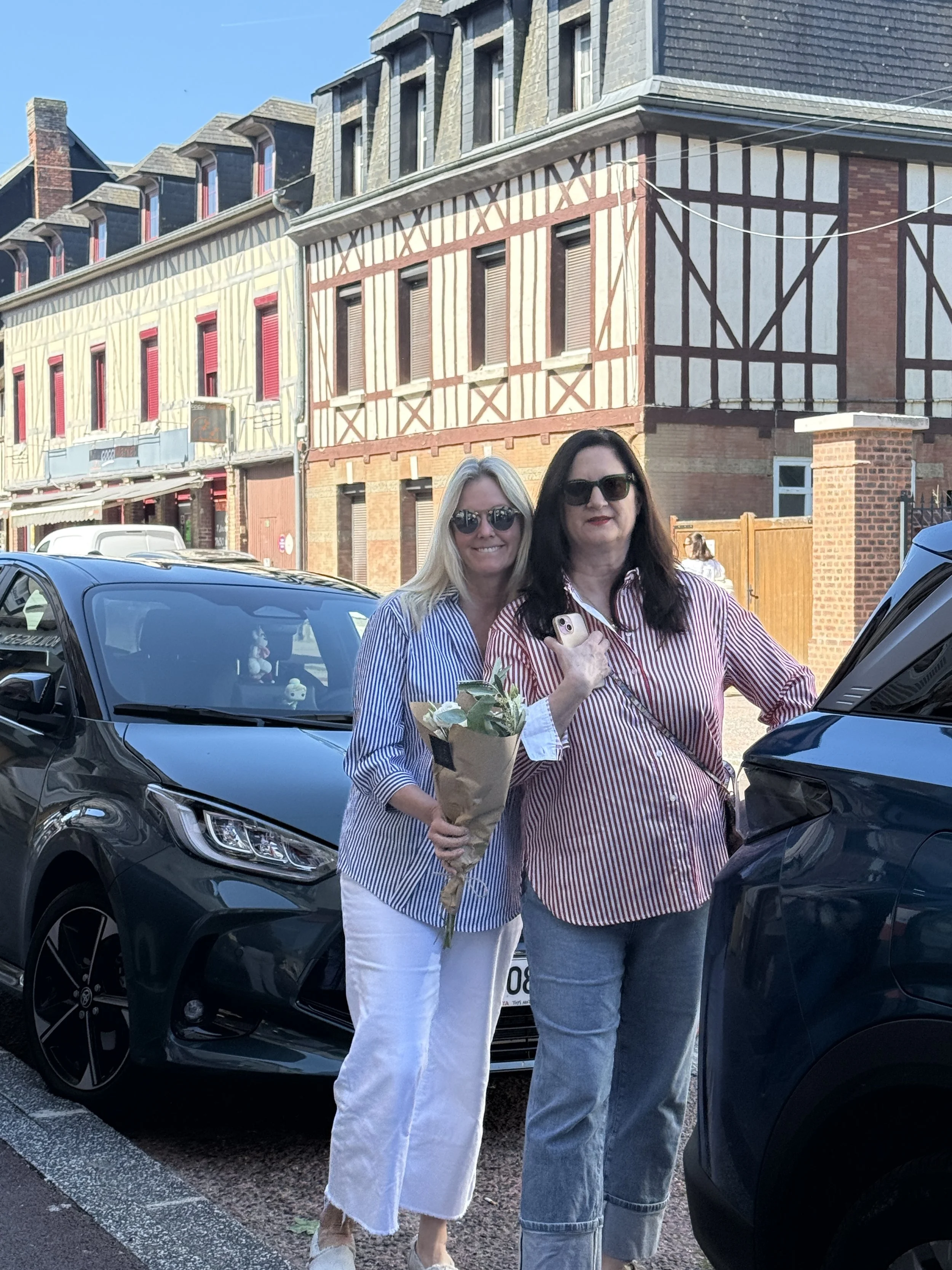 Two women standing between parked cars in front of a historic half-timbered building, one holding a bouquet of flowers and smiling at the camera.