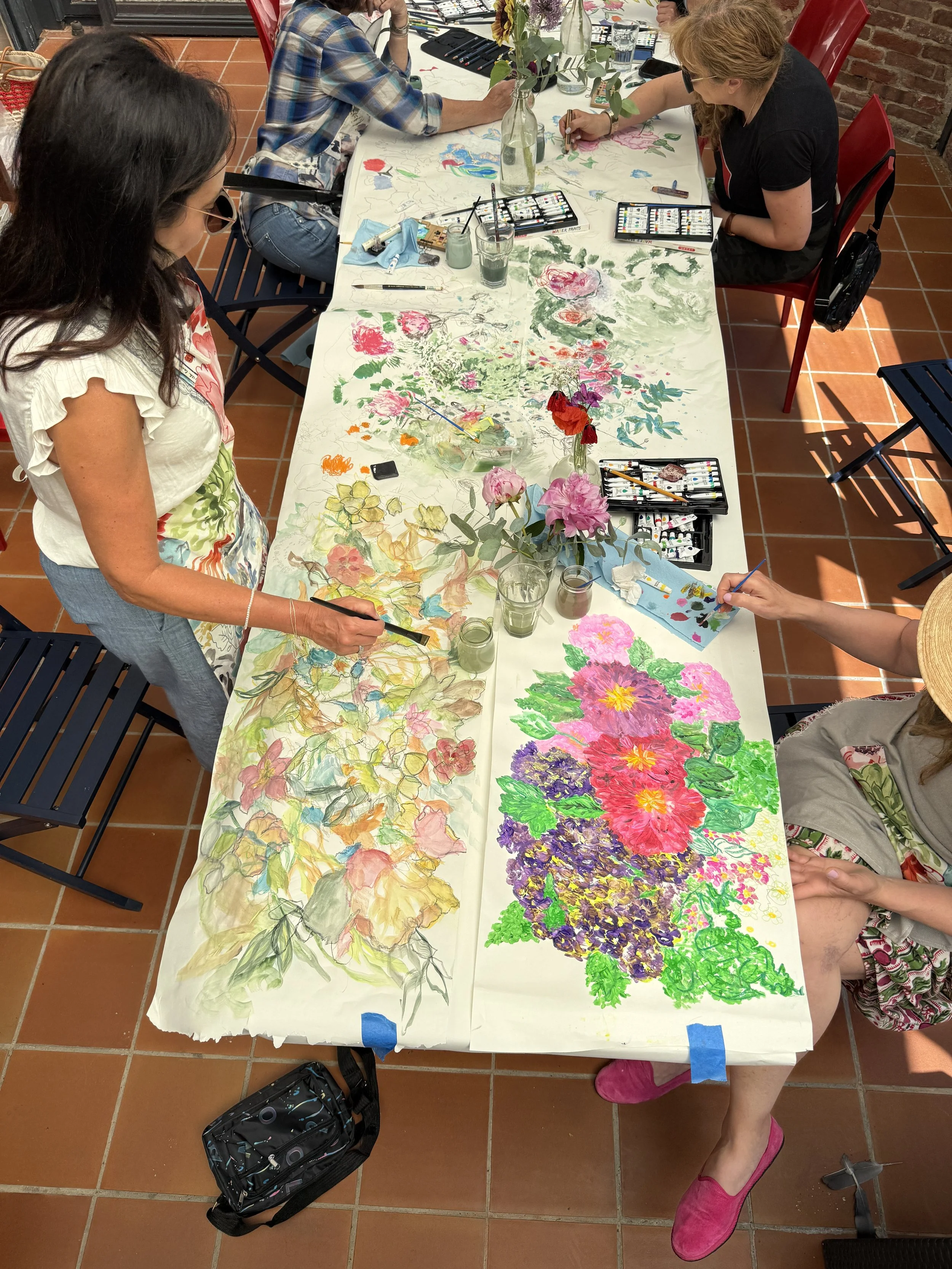 People sitting around a table painting floral watercolor art on large sheets of paper, with art supplies and glasses of water on the table.