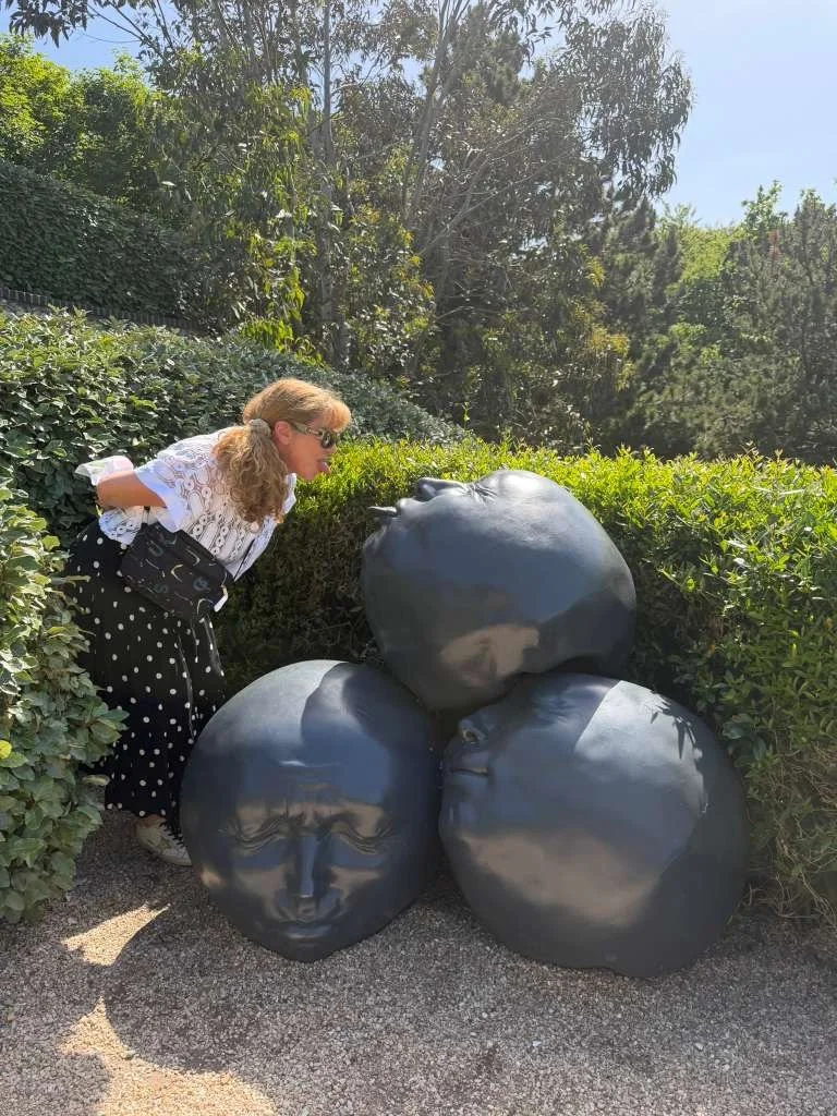 A woman with curly hair, wearing sunglasses and a polka dot skirt, leans in to look closely at four black sculptures resembling human faces, placed among green bushes and trees on a sunny day.