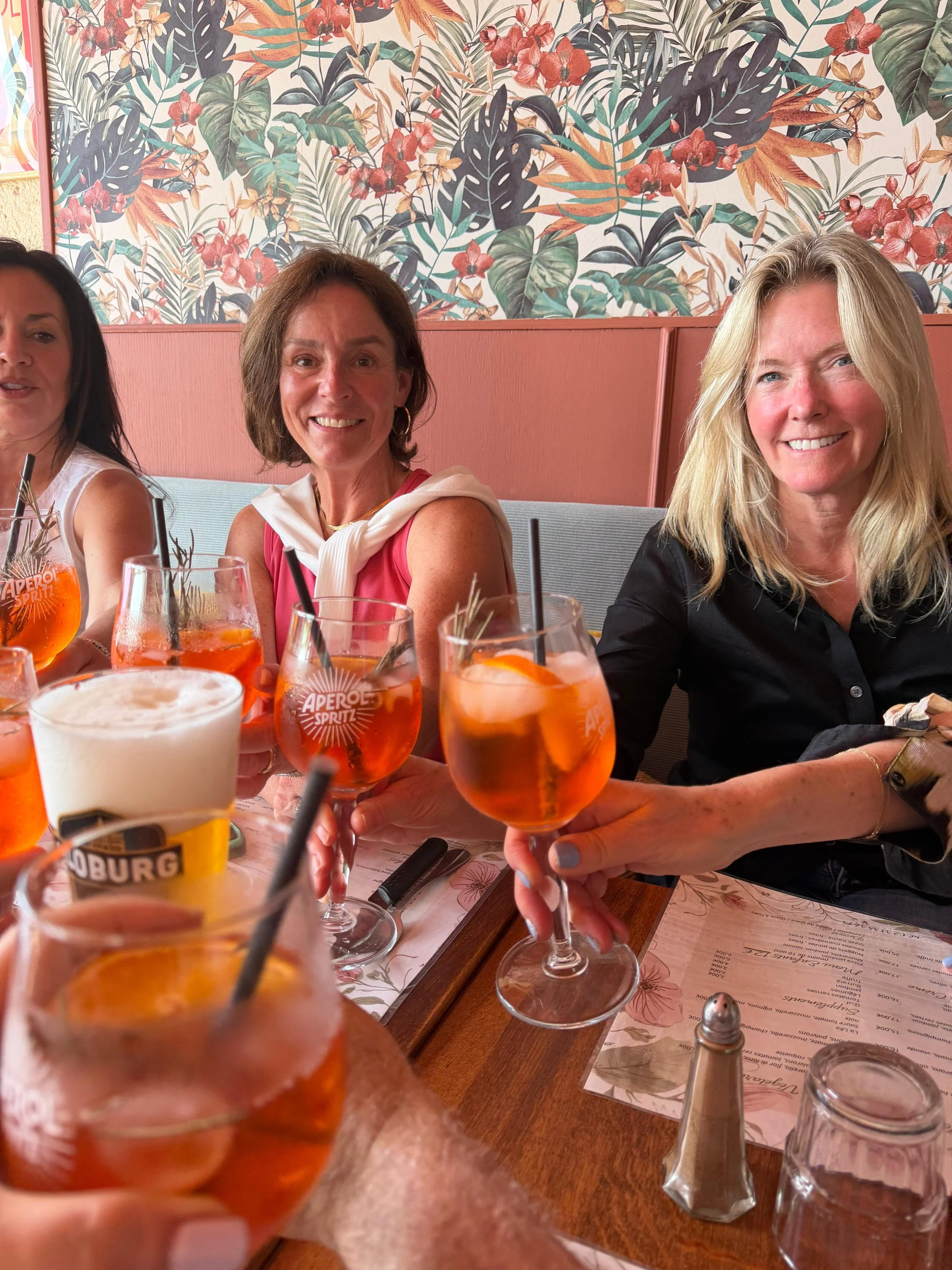 A group of women sitting at a table in a restaurant, holding glasses of spritz cocktails with orange slices, smiling, with a floral wallpaper in the background.