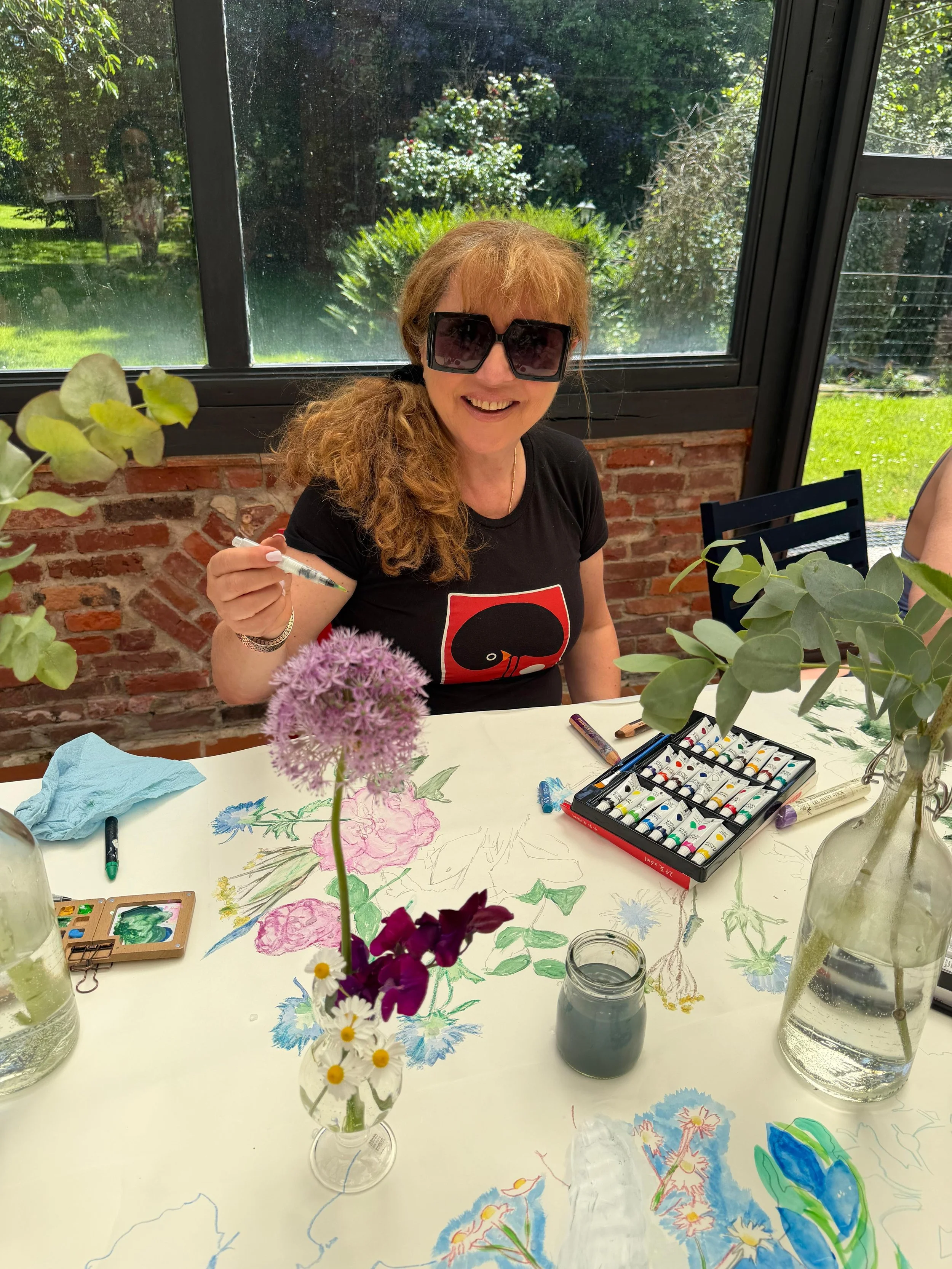 A woman wearing large sunglasses sitting at a table with floral drawings and watercolor paints, holding a pencil, with plants and a brick wall outside the window behind her.