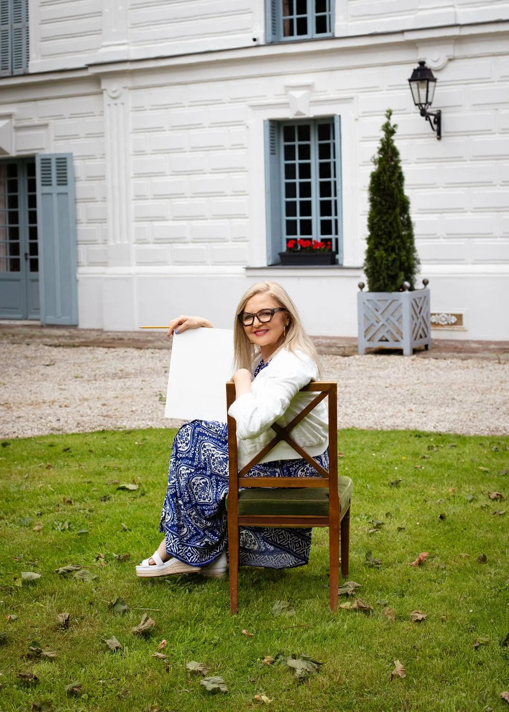 A woman with glasses and blonde hair sitting on a wooden chair in a garden, holding a white notebook and pencil, in front of a white house with blue shutters and a potted plant.
