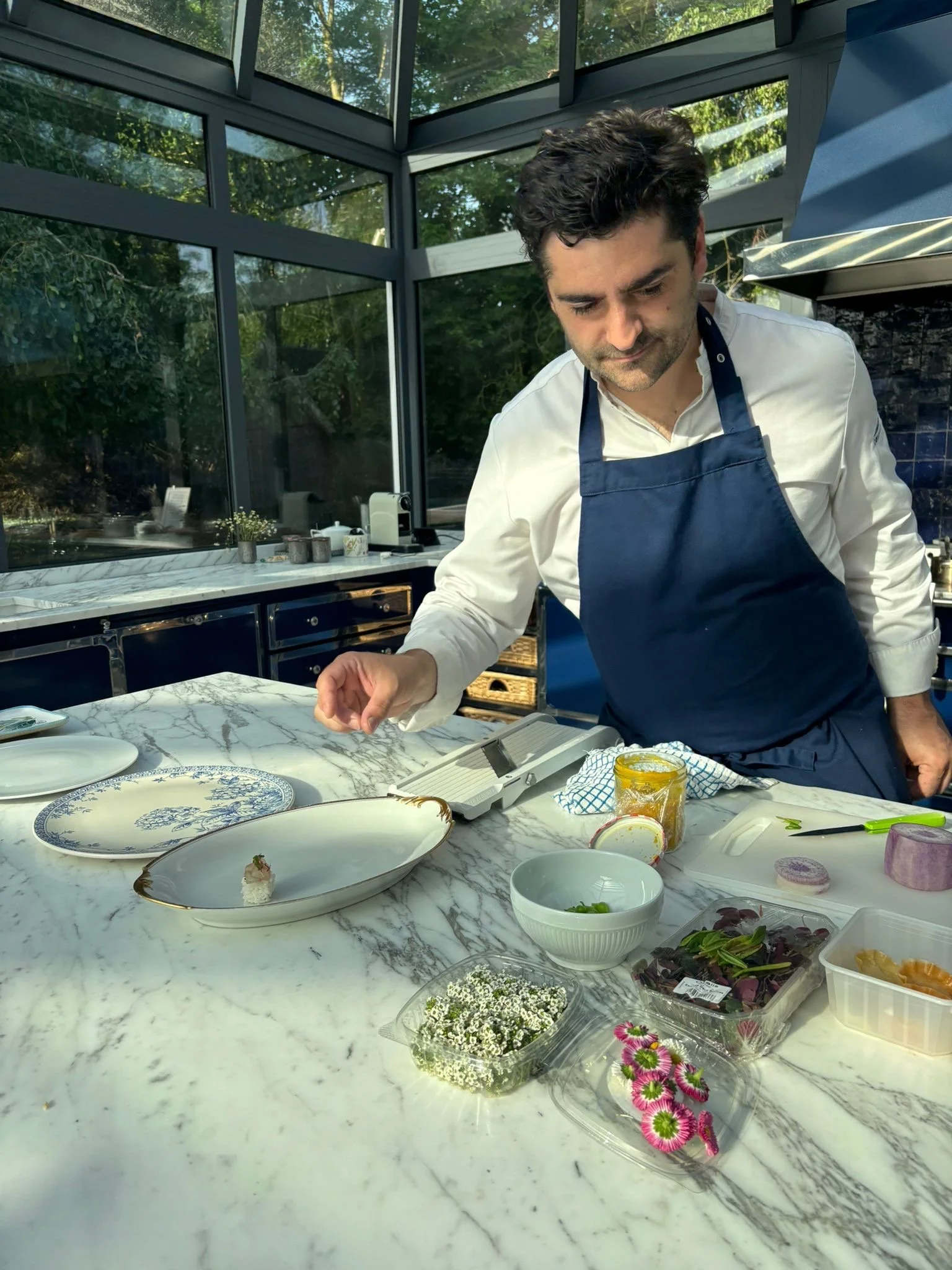 A man wearing a navy apron and a white shirt standing at a marble kitchen counter with various dishes, bowls, and ingredients, in a bright kitchen with large glass windows and green trees outside.