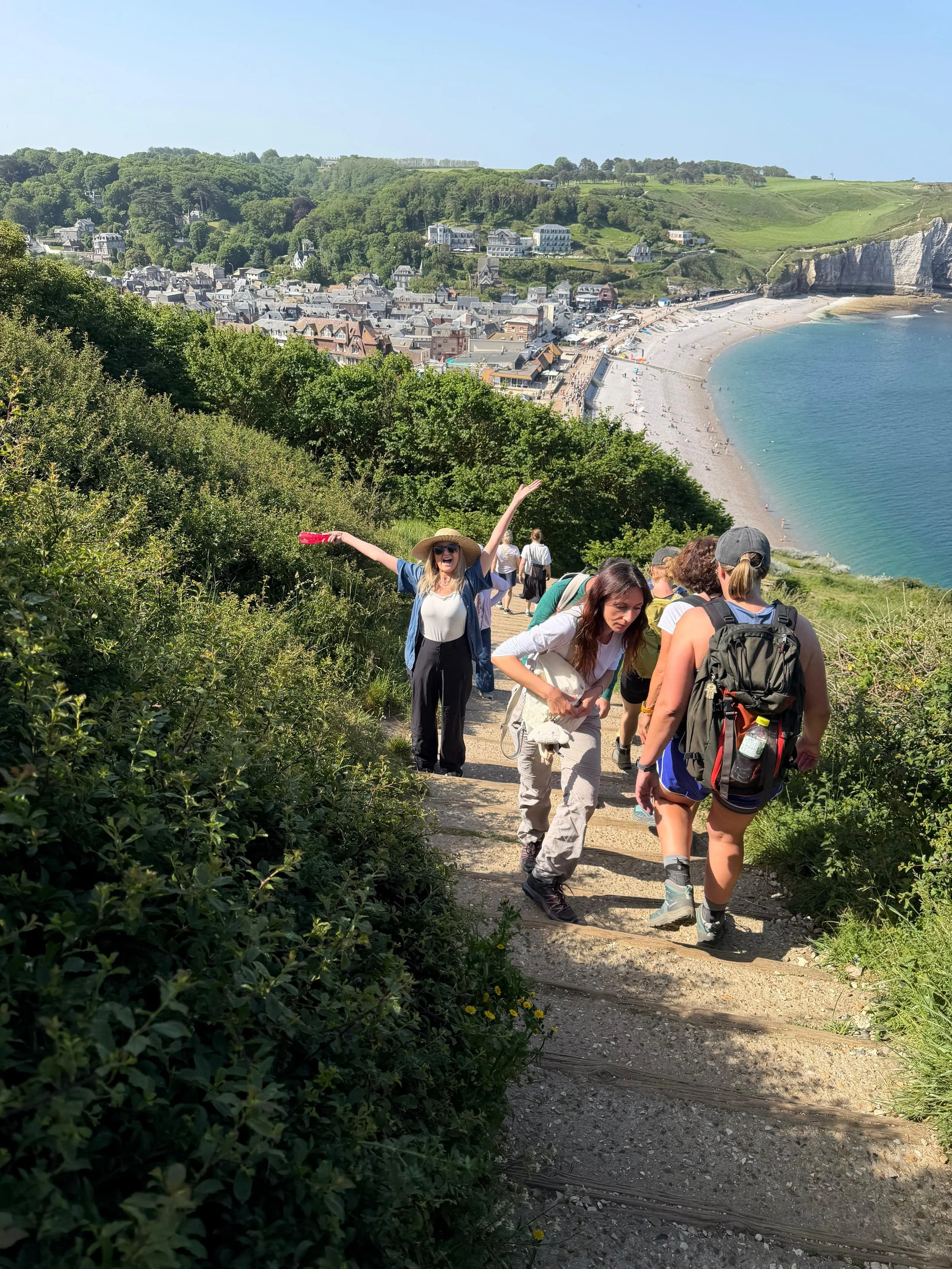 Group of hikers walking on a trail along a hillside overlooking a coastal town and beach with cliffs and blue ocean in the background.