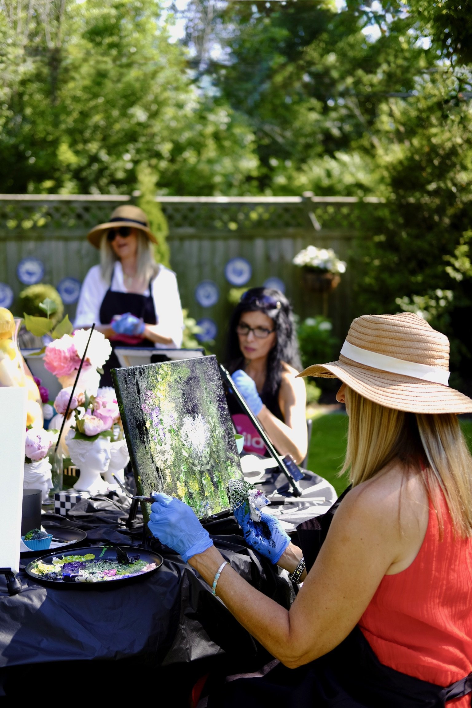 A woman with long blonde hair wearing a straw hat and red sleeveless top is painting on a canvas at a outdoor art event. There are friends and art displays in the background surrounded by green trees.