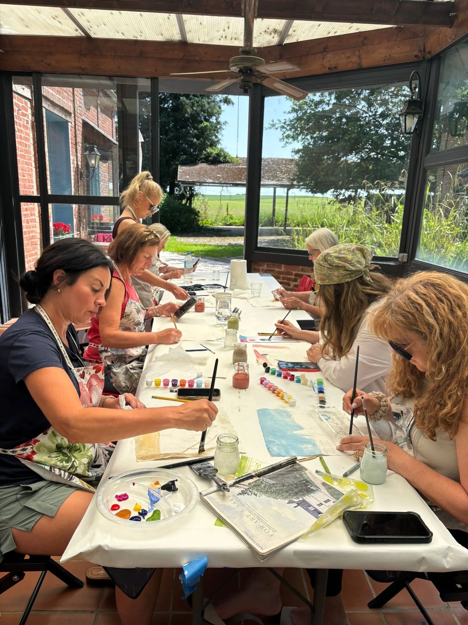 Several women sit around a long table painting with watercolor paints on paper. The table has paints, brushes, a sketchbook, and water jars. Some women are wearing aprons and sunglasses. The room has large windows showing a sunny outdoor scene with trees and buildings.
