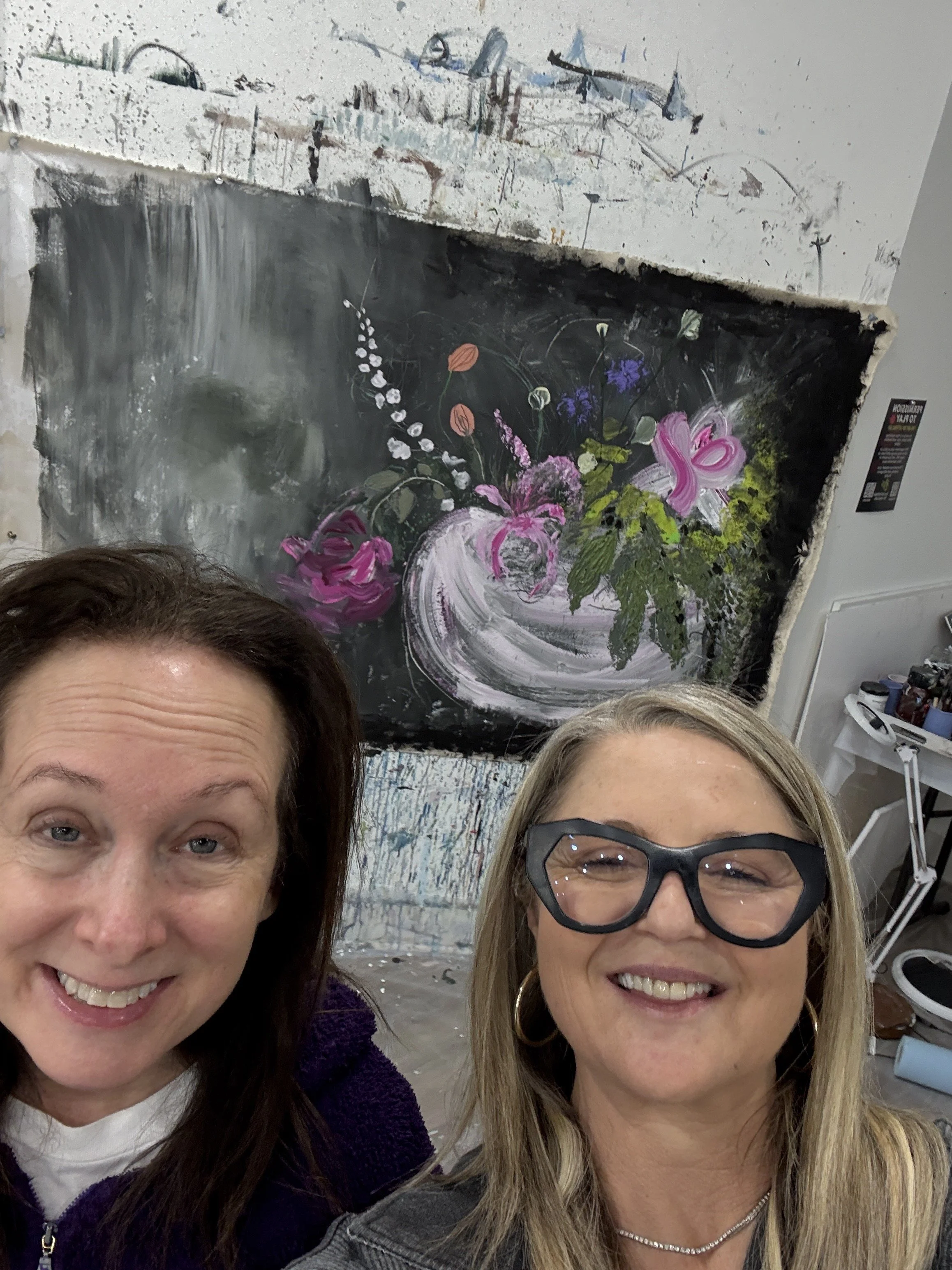 Two smiling women taking a selfie in front of a large, colorful flower painting at an art studio.