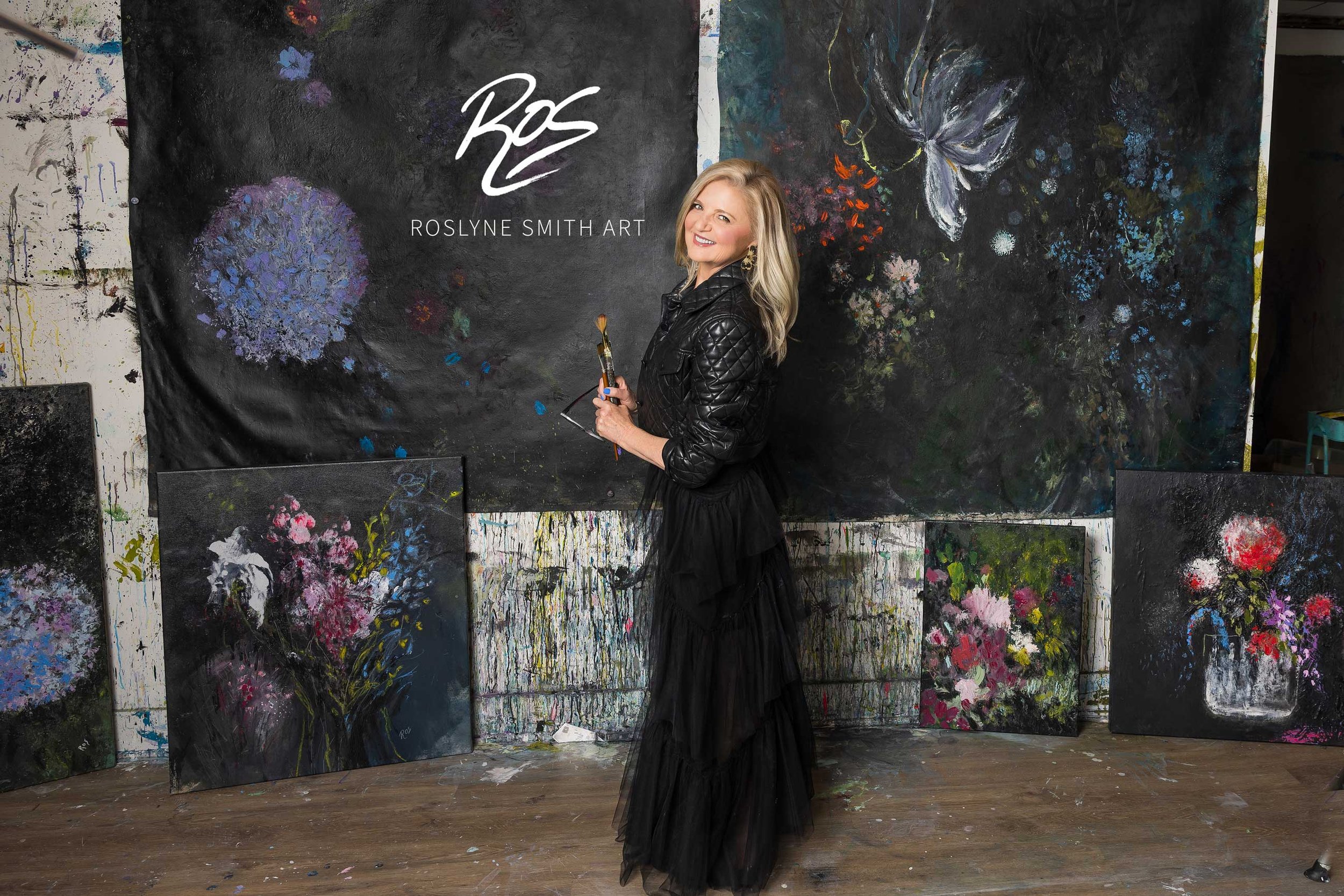 A woman with blonde hair wearing a black quilted jacket and a long black skirt, smiling and holding a paintbrush, standing in front of a large black background wall with colorful abstract floral and butterfly paintings, and the text 'Roslyne Smith Art' with a signature above.