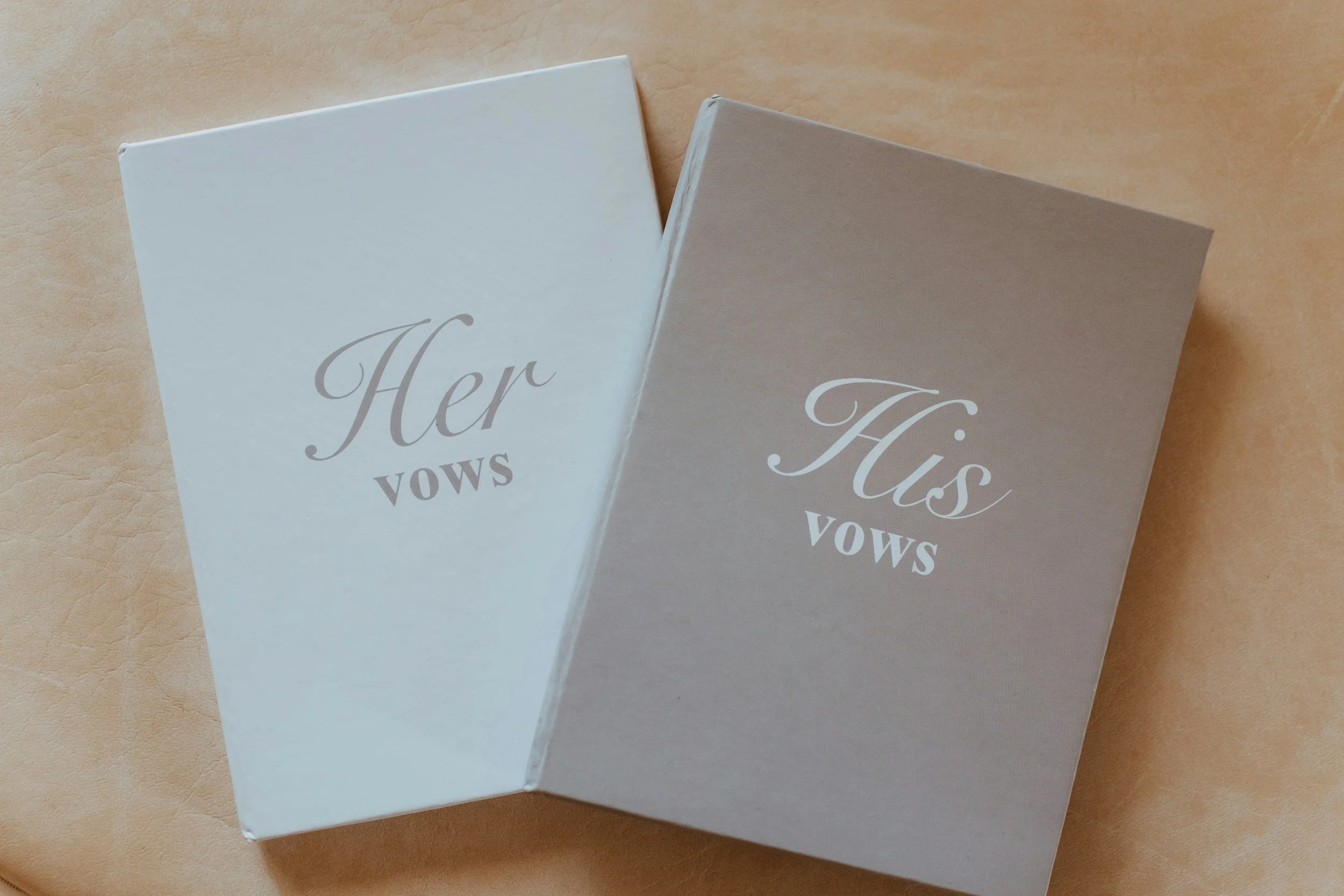 Two greeting cards on a tan surface, one white and one light gray, both with the words "Her Vows" and "His Vows" written in elegant script.