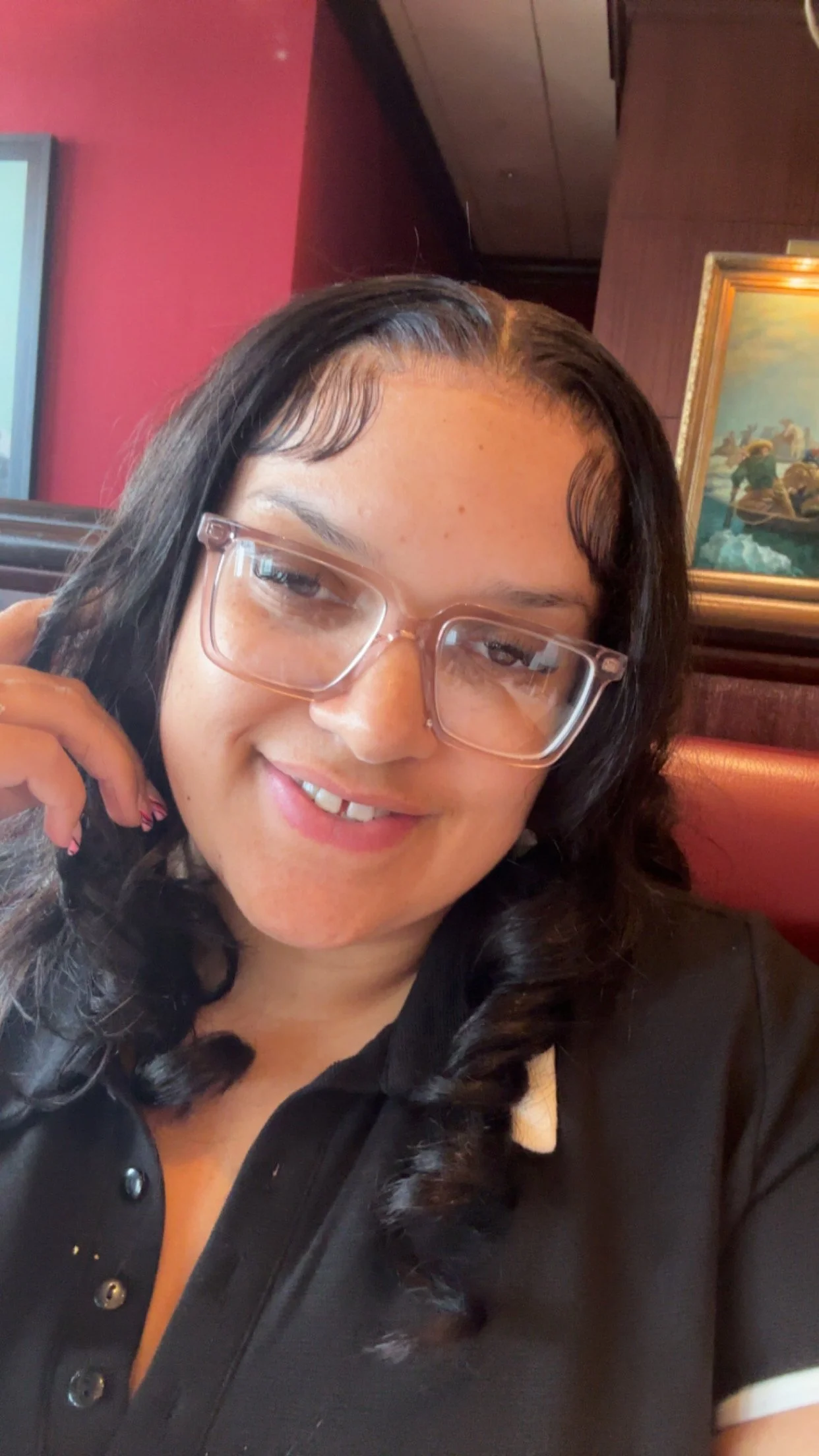 A woman with long dark hair, glasses, and a light smile, sitting in a restaurant with a framed picture of a landscape on the wall behind her.