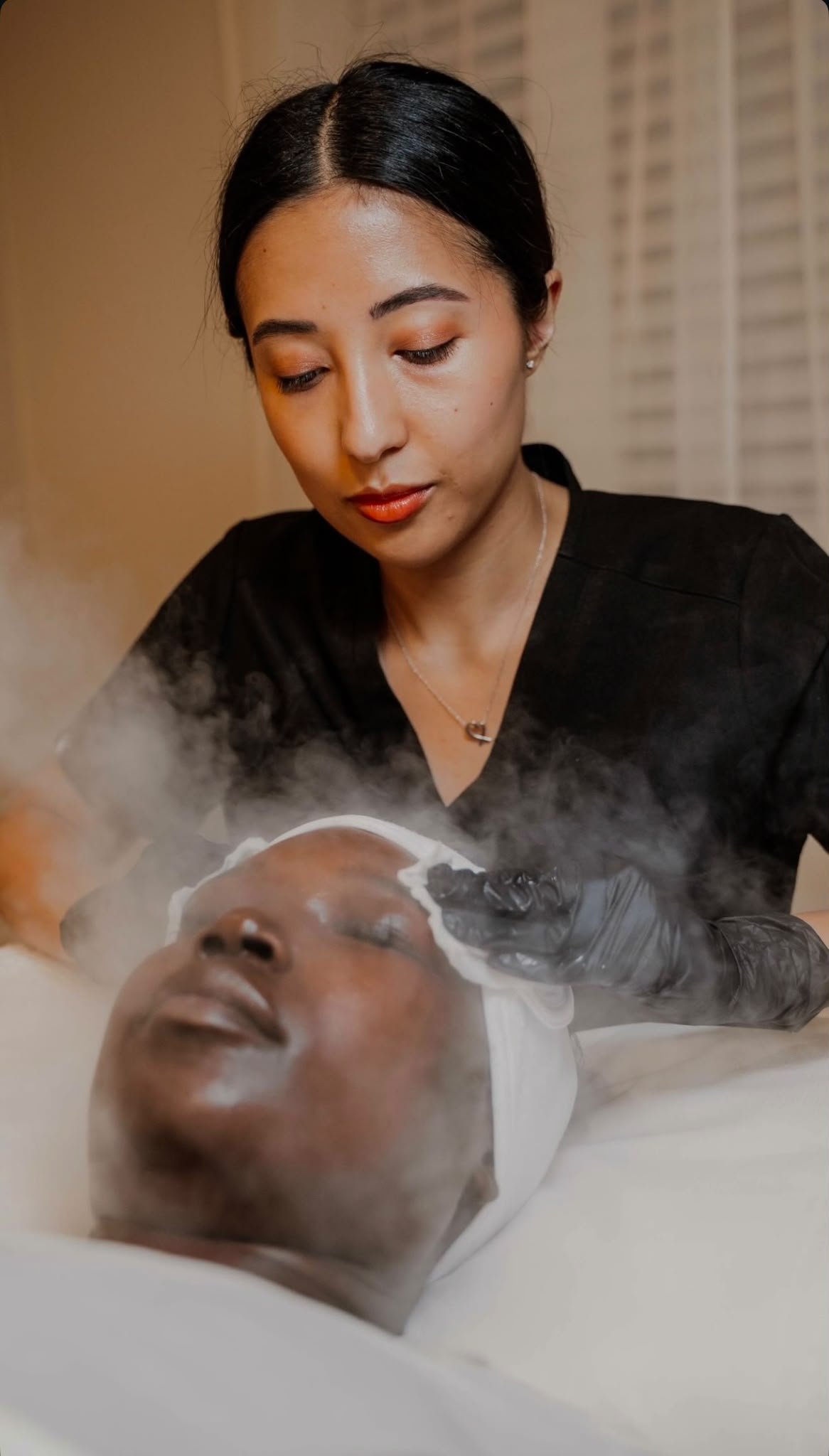 A woman with dark hair and earrings performing a facial treatment on a client lying with closed eyes, using a white sheet or cloth on their face. The woman is focused, wearing a black top, in a room with warm lighting.