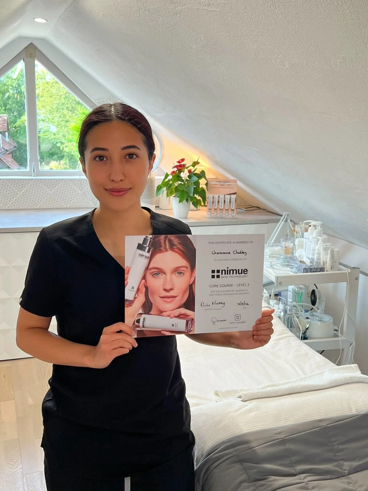 A woman smiling and holding a certificate of completion for Nimue Skin Technology, standing in a skincare or medical clinic with treatment supplies and a bed in the background.