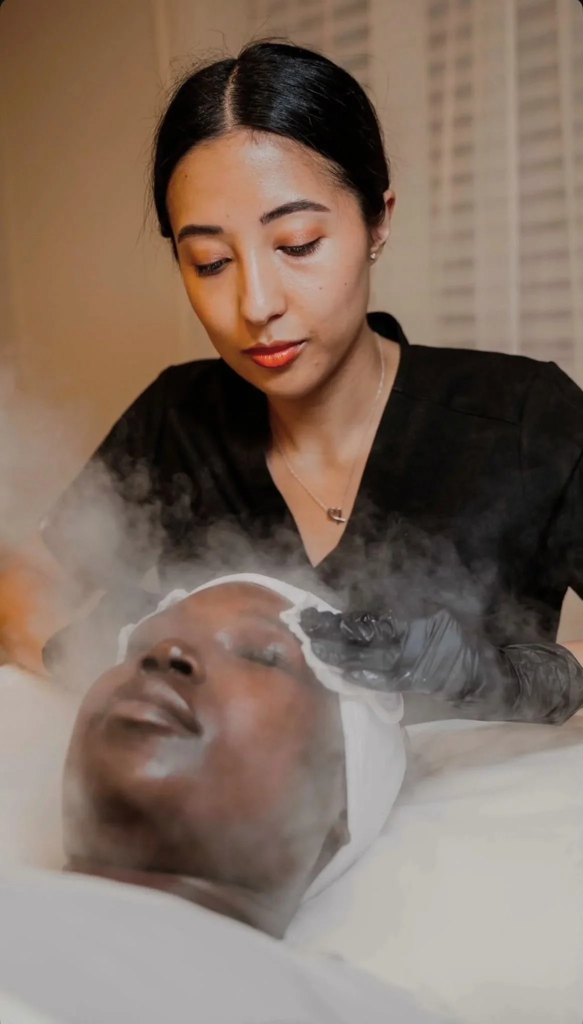 A woman with dark hair and skin tone performing a facial treatment on a woman lying down. The woman lying down has closed eyes, and the woman performing the treatment is focused and wearing a black top and gloves, with steam or vapor visible around them.