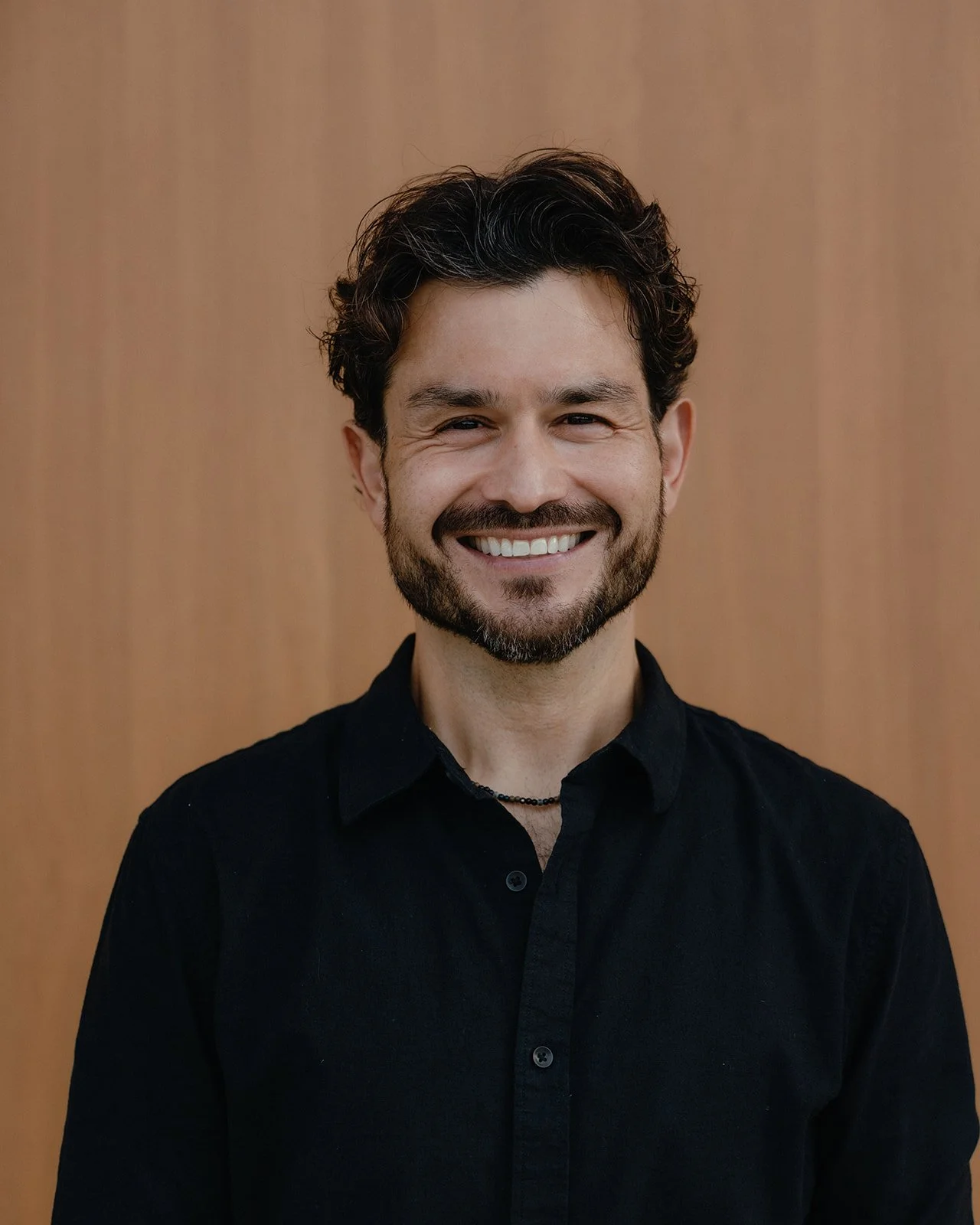 Smiling man with dark curly hair and beard, wearing a black shirt, standing against a brown wooden background.