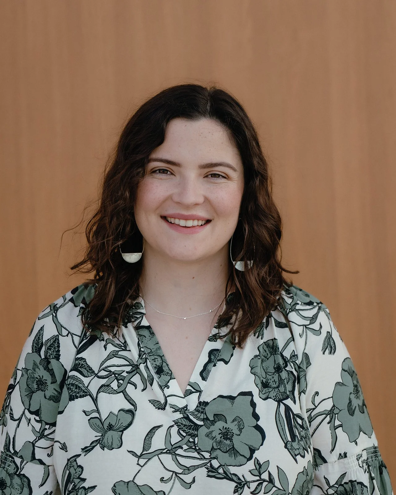 A woman with curly dark hair and fair skin, smiling, wearing a floral blouse with black and white pattern, and earrings, standing against a wooden background.