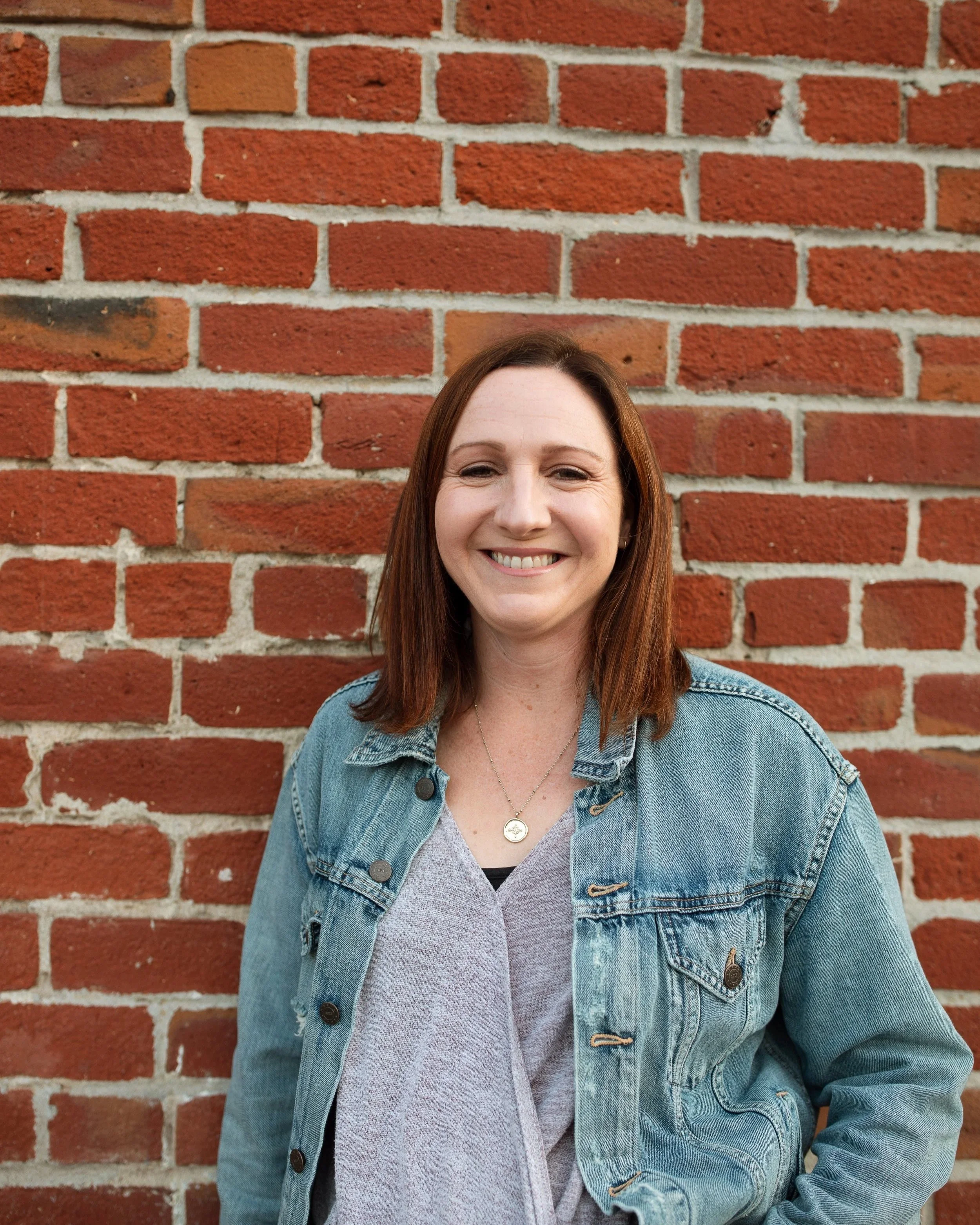 Smiling woman with brown hair wearing a gray shirt and a denim jacket standing in front of a red brick wall.