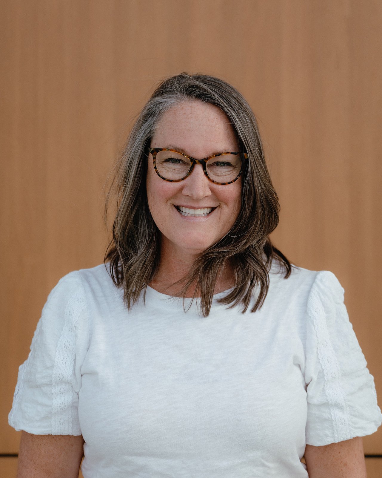 A woman with shoulder-length brown hair wearing glasses and a white top smiling in front of a wooden background.