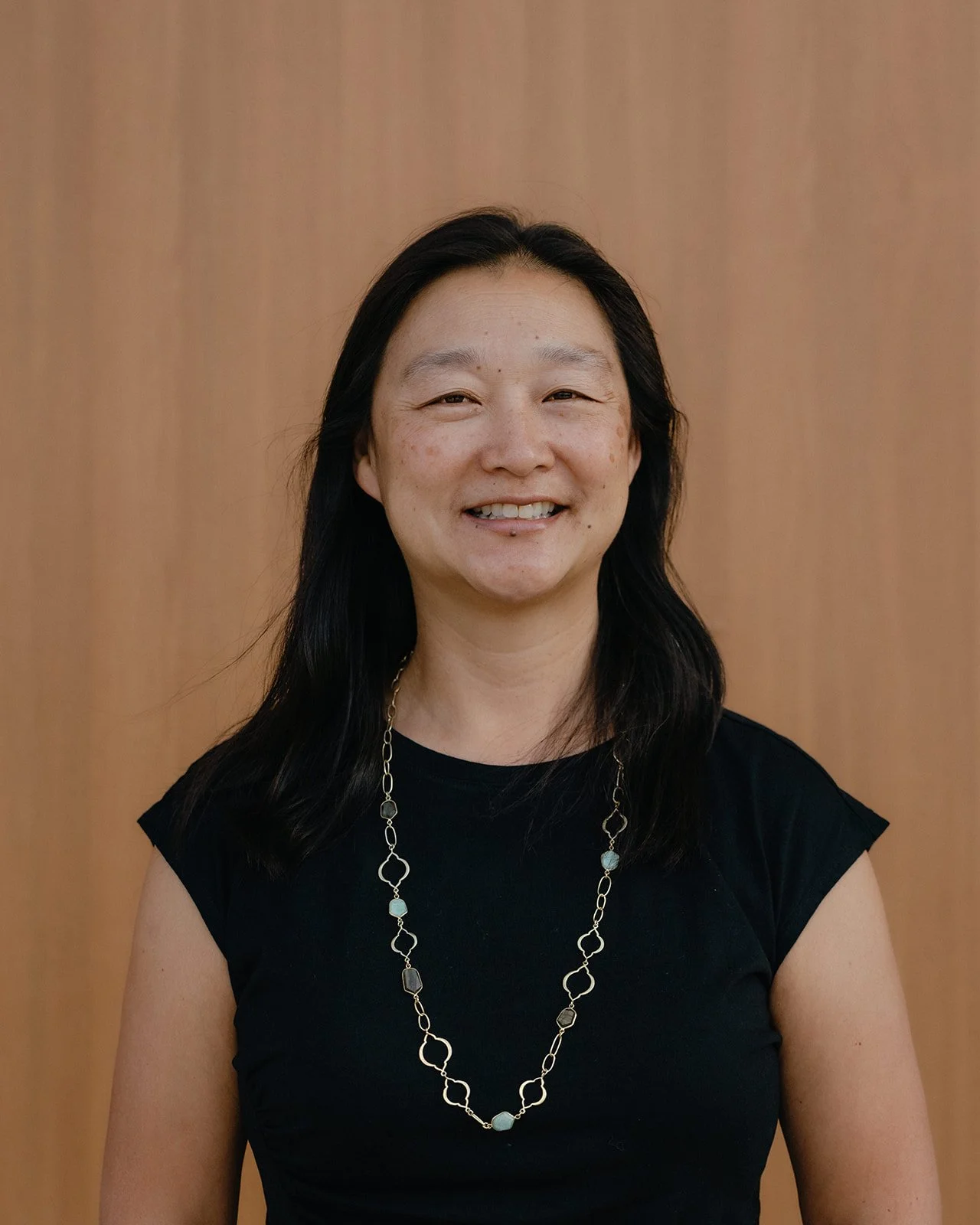 Portrait of a woman with black hair wearing a black top and a silver necklace, standing in front of a wooden background.