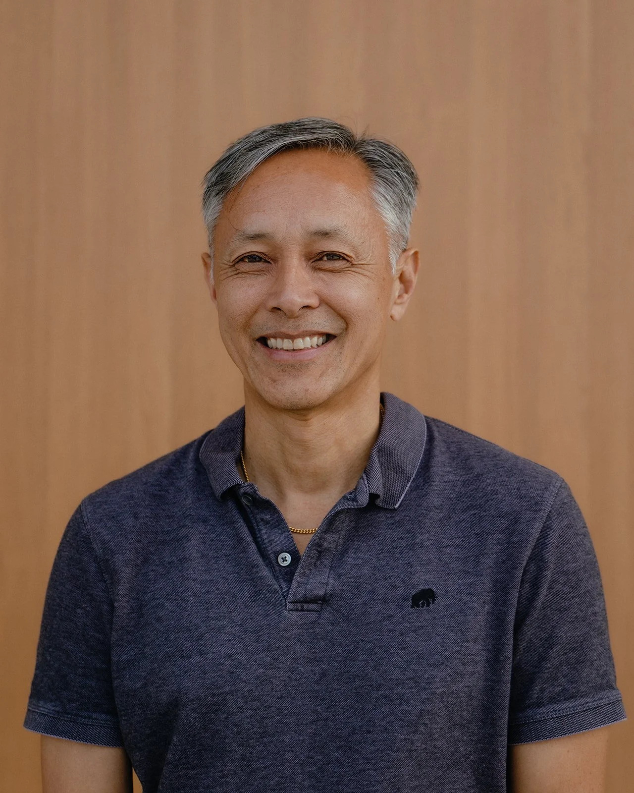 A middle-aged man with gray hair smiling, wearing a dark blue polo shirt with a small bear logo, standing against a wooden background.
