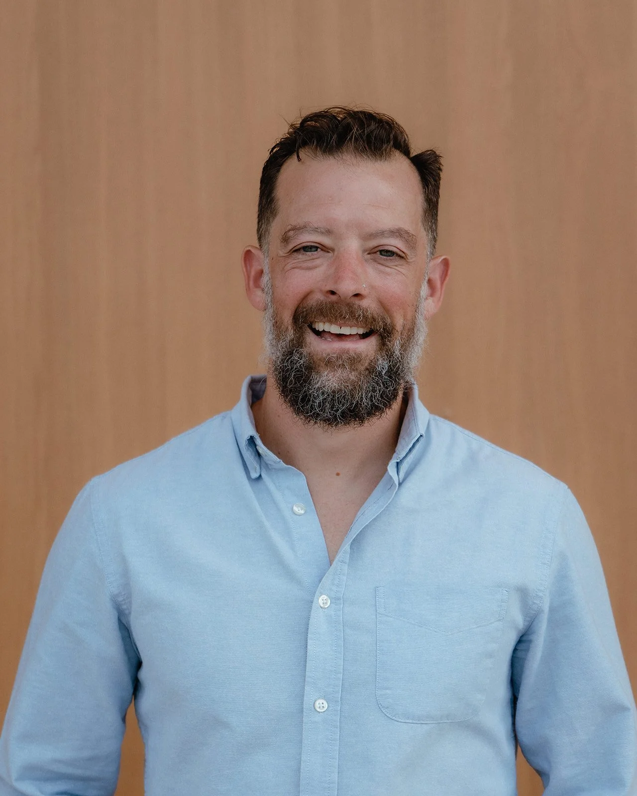 A smiling man with a beard, wearing a light blue button-up shirt, standing in front of a wooden background.