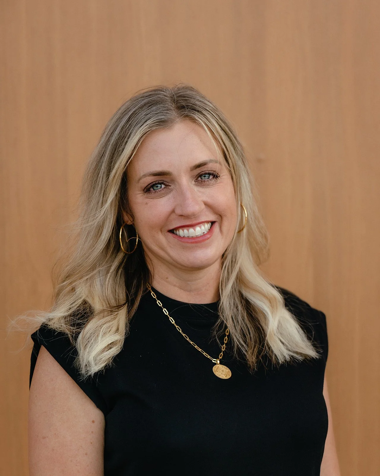 A smiling woman with long blonde hair, blue eyes, and gold hoop earrings, wearing a black top with gold jewelry, standing against a wooden background.