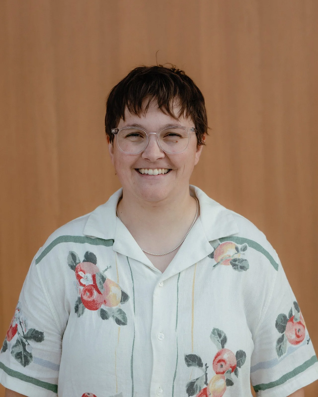 A person with short dark hair and glasses smiling, wearing a white shirt with a floral pattern, standing against a wooden background.