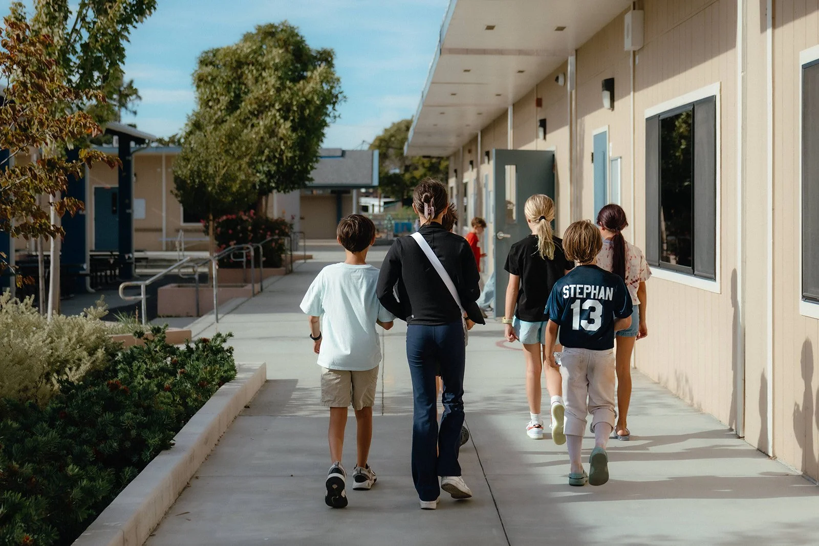 A group of children and a woman walk down an outdoor sidewalk outside a school building on a sunny day.