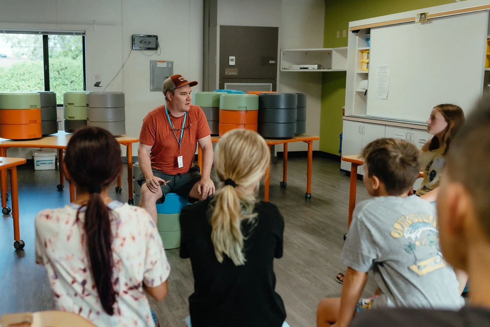 Young man giving a presentation to children in a classroom, sitting on a blue ottoman, with storage bins and a whiteboard behind him.