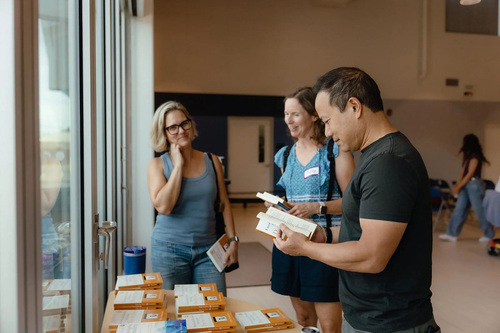 Three people at a church service, looking at books, with a table of other printed materials. The setting appears to be indoors with a casual atmosphere.