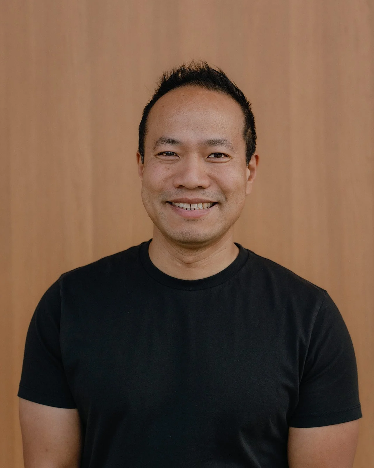 A man with short black hair, smiling, wearing a black t-shirt, standing in front of a wooden background.
