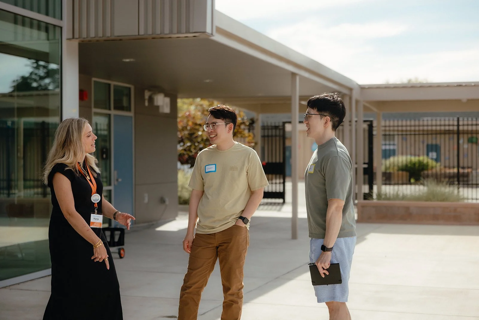 Three people, two young men and a woman, are standing outside having a conversation, smiling and laughing. The woman is wearing a black dress and an orange lanyard. The young men are wearing casual clothes and glasses, one with a name tag that says "Simon." It is daytime with a building, trees, and a fence in the background.