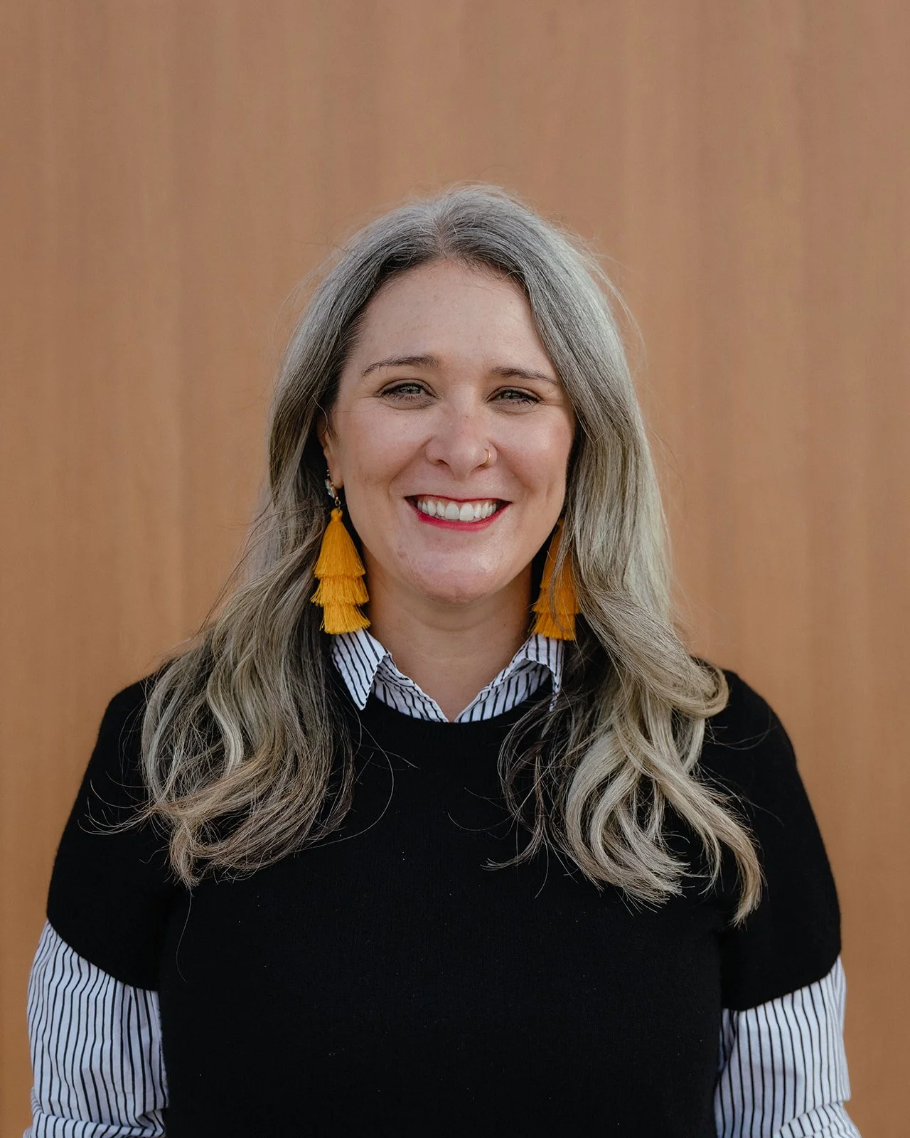 A woman with long gray hair, smiling, wearing a black sweater, a striped collared shirt, and yellow tassel earrings, standing in front of a plain wooden background.