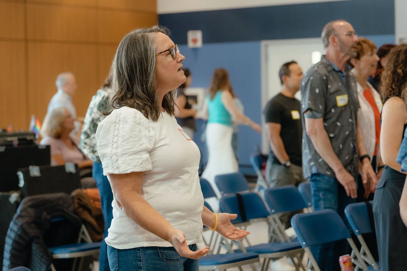 A woman with glasses, wearing a white blouse and jeans, standing with her hands open in a crowded church service.