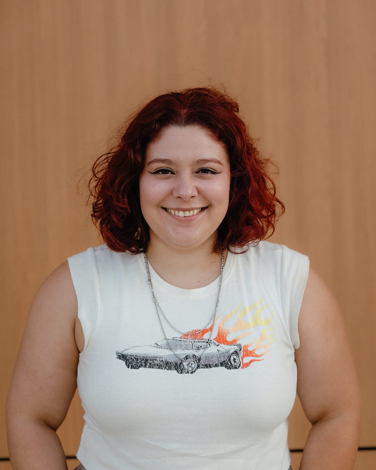 A young person with red, curly hair smiling, wearing a sleeveless white t-shirt with a car and flame graphic and a silver necklace, standing in front of a wooden background.