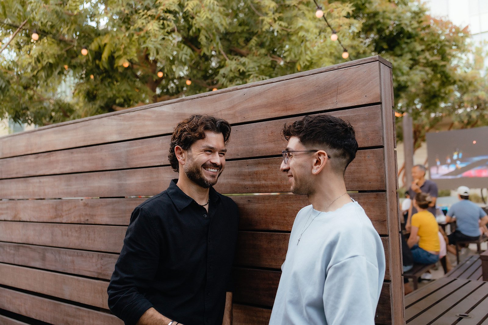 Two men smiling and talking to each other while standing behind a wooden privacy screen outdoors, with trees and people sitting at tables in the background.