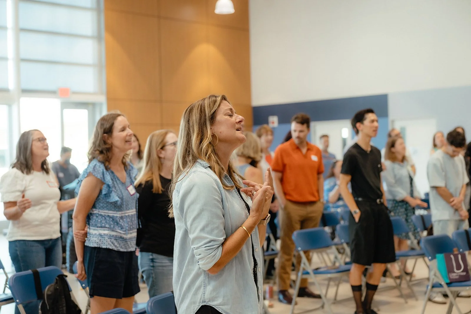 A group of people standing with their eyes closed and hands over their hearts, engaged in a moment of prayer or reflection in a well-lit, modern conference room with wooden and white walls.