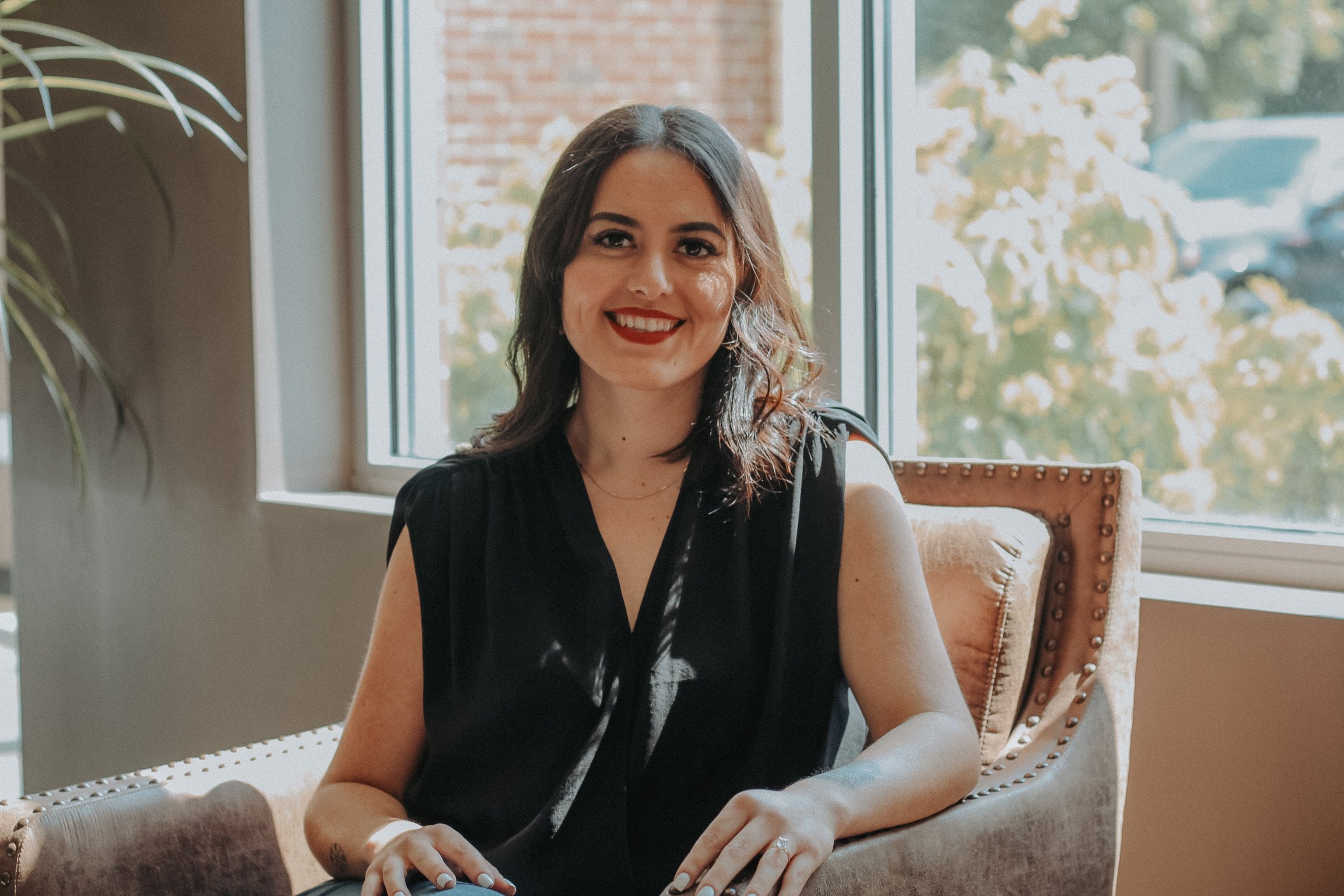 A woman with dark brown hair and red lipstick sitting on an upholstered armchair, smiling at the camera, with a large window behind her showing trees and a blue sky.