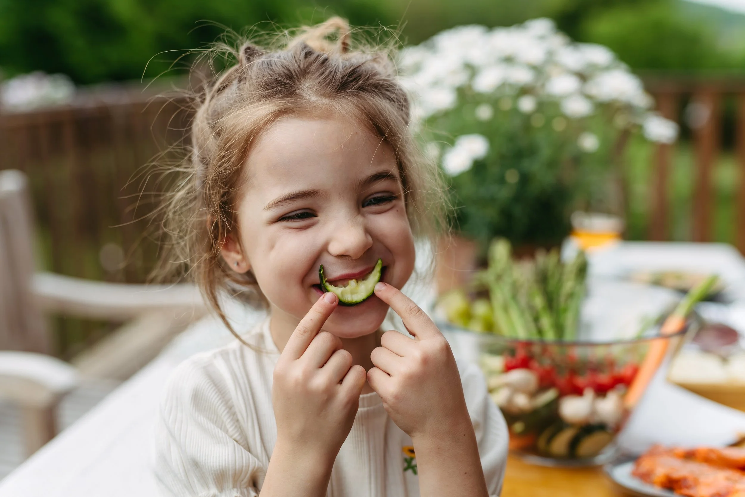 A young girl with light brown hair smiling and biting into a slice of cucumber outdoors in a garden or patio setting, with a table filled with various salads and food in the background.