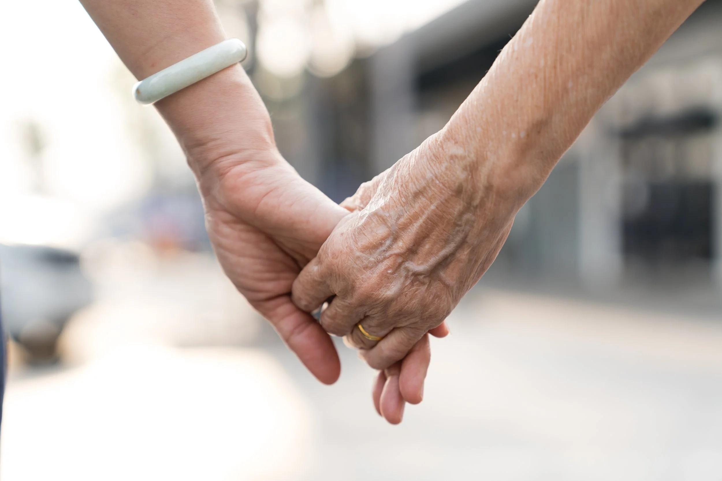 Close-up of two elderly hands holding each other, one hand wearing a white bracelet, with blurred background.