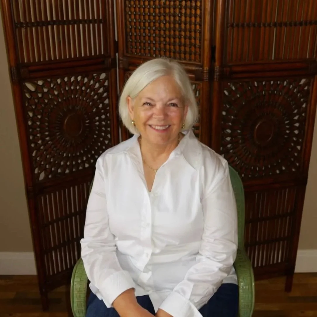 An elderly woman with short white hair smiling, sitting on a green chair in front of a decorative wooden room divider.