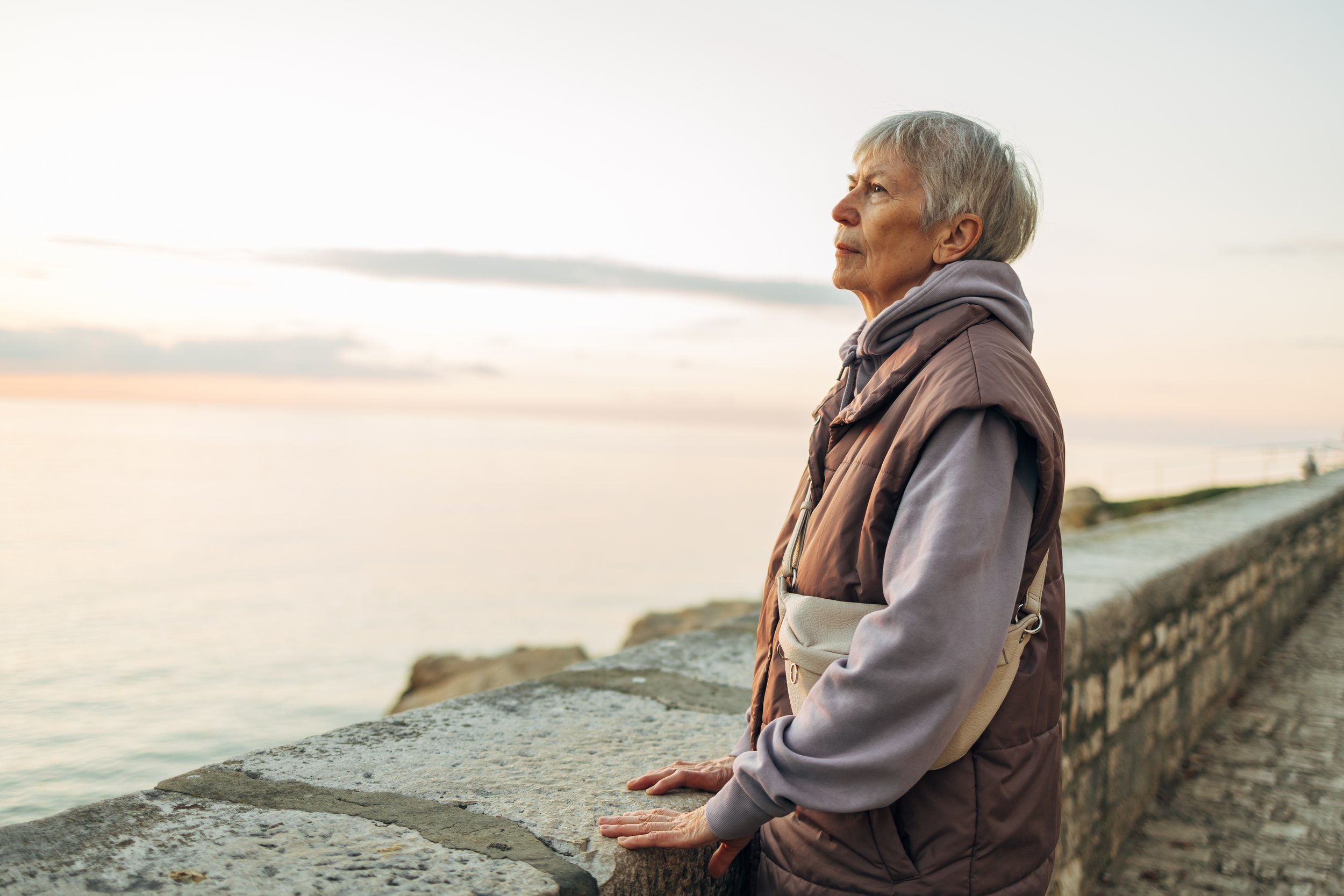 Elderly woman gazing at the ocean while standing at a stone railing during sunset, wearing a hoodie and vest.